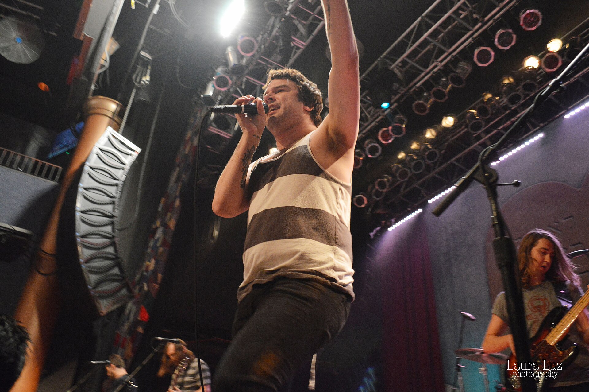 Max Bemis sings into a handheld mic with one arm thrown up, in a striped vest, shot from below against a rig of stage lights.