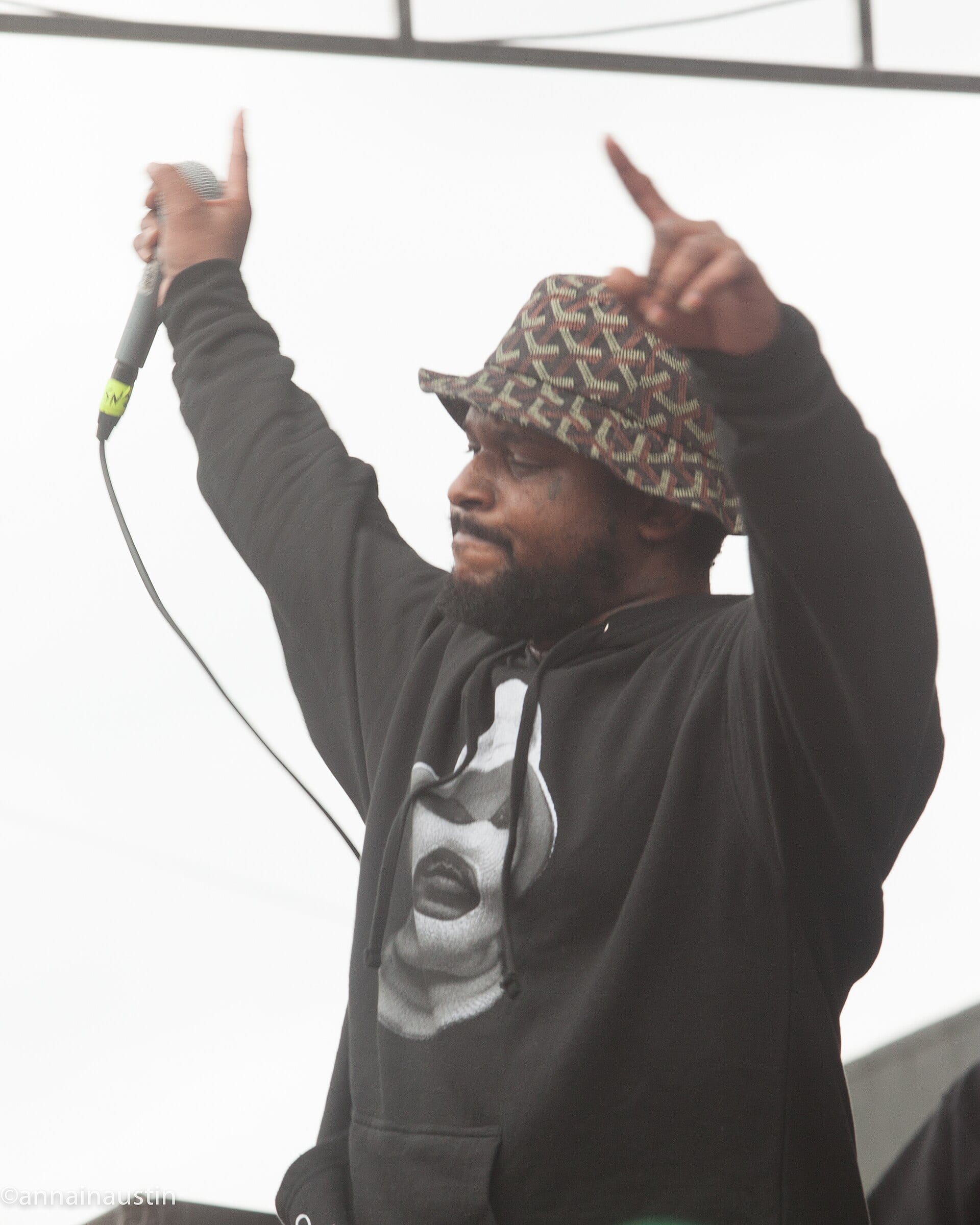 ScHoolboy Q raises both arms and holds a microphone while performing on an outdoor stage, wearing a patterned bucket hat and a hoodie.