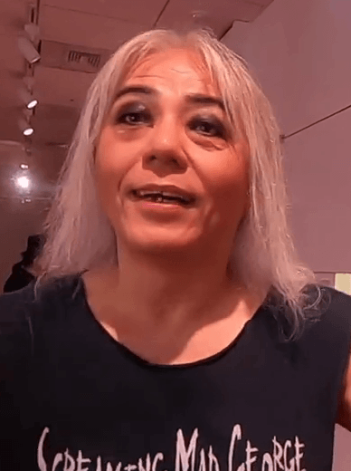 Screaming Mad George, with long gray hair, speaks at his art retrospective wearing a black t-shirt printed with his own name.