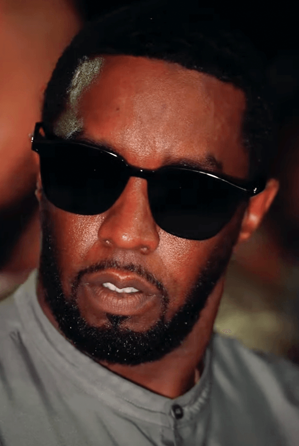 Sean Combs, wearing dark sunglasses and a grey jacket, speaks during a close-up interview shot.
