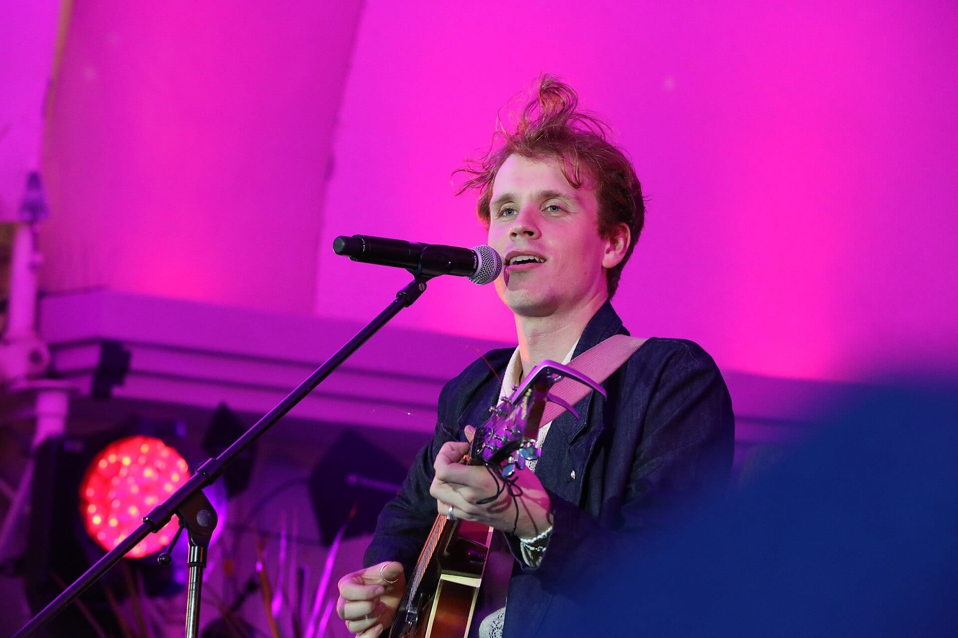 Sebastian Schub sings into a microphone while playing an acoustic guitar on stage, his hair tousled, lit by pink and purple stage light.