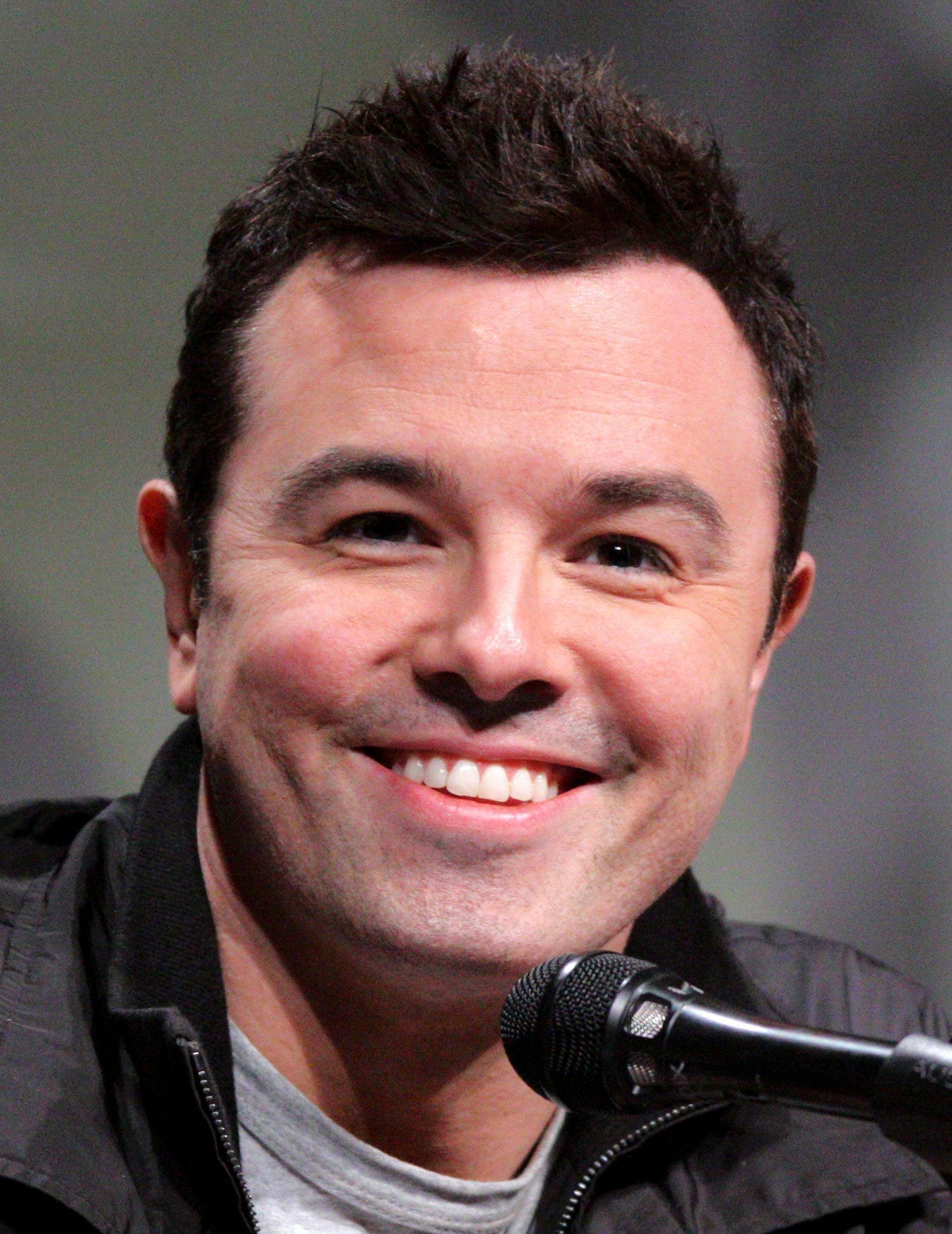 Seth MacFarlane smiles at a close angle into a microphone, wearing a dark zip-up jacket over a gray T-shirt.