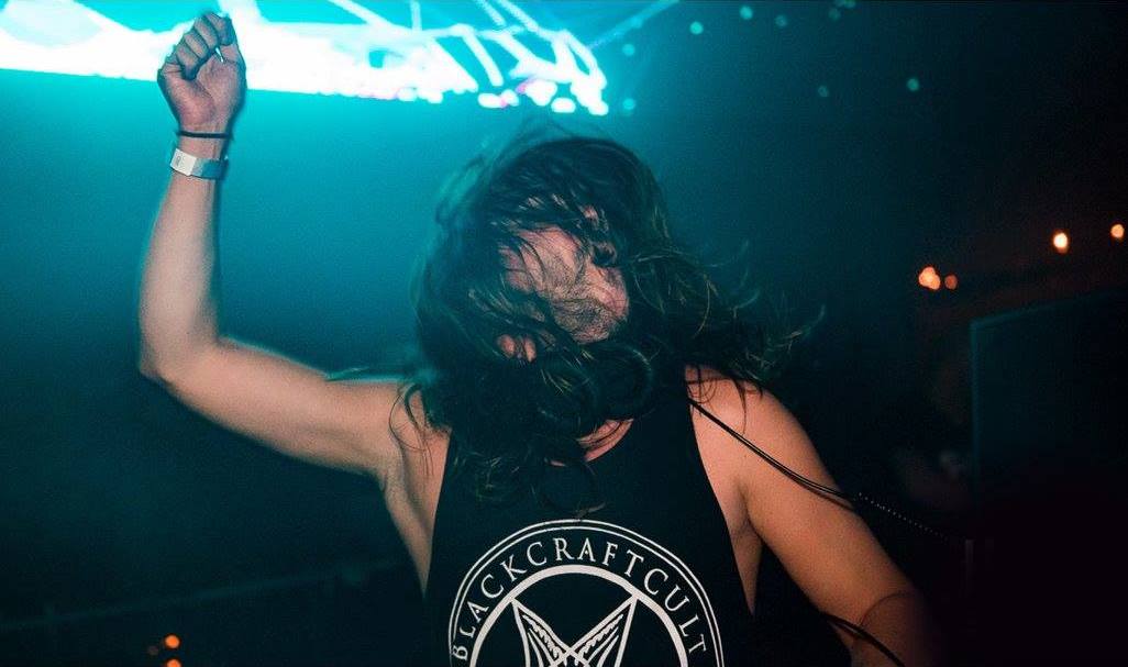 Seven Lions throws his head forward behind the decks in a black sleeveless top, long hair covering his face, in white and violet light.