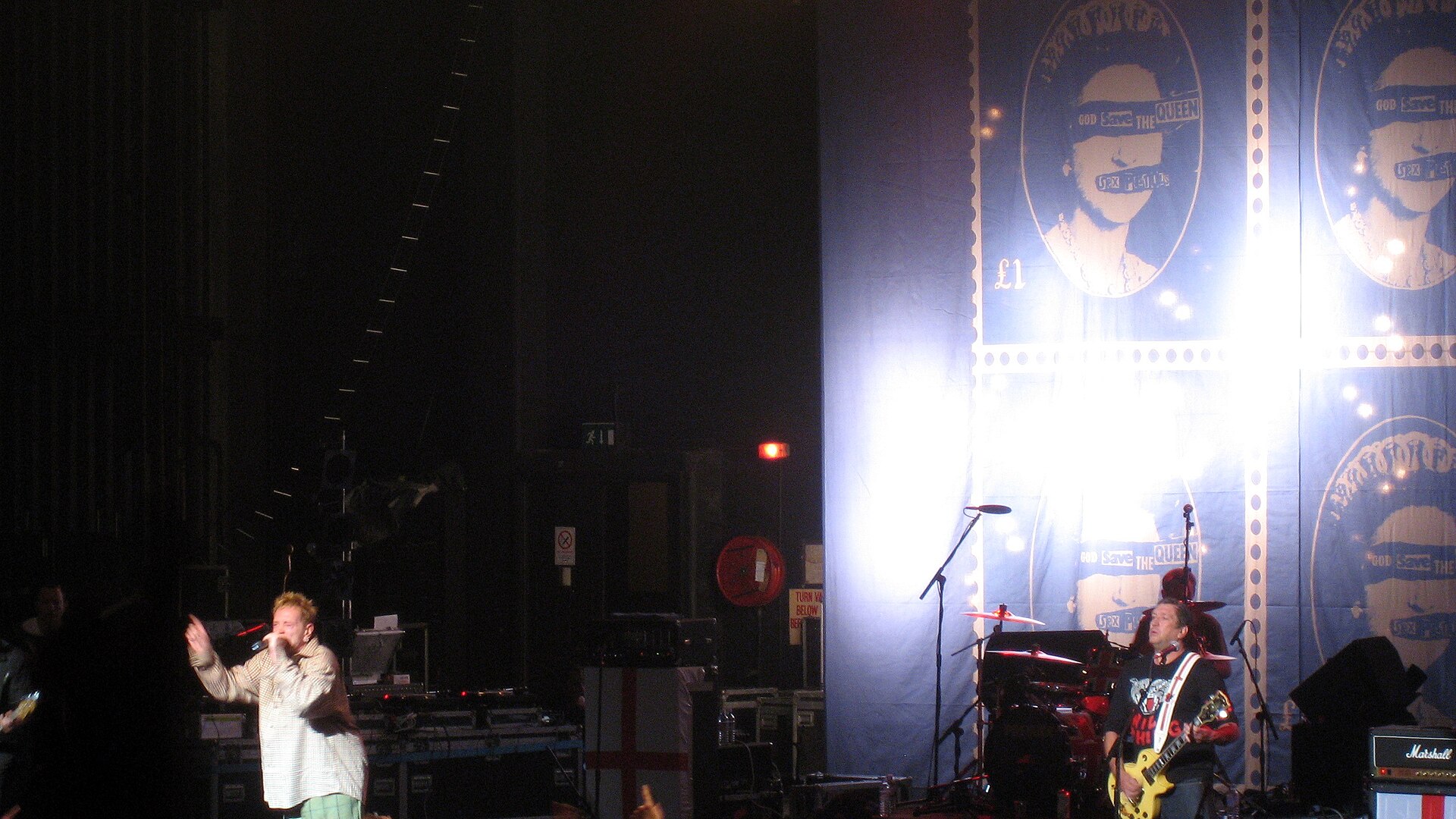 Two members of the Sex Pistols on a wide stage, a singer at left with a microphone and a guitarist at right, in front of a giant God Save the Queen postage-stamp backdrop.