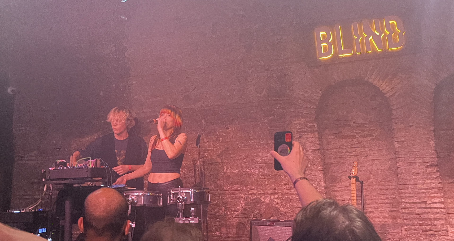 Sextile play a smoky brick-walled club under a neon Blind sign, one member singing into a handheld microphone over electronic drum pads while the other works a table of hardware.