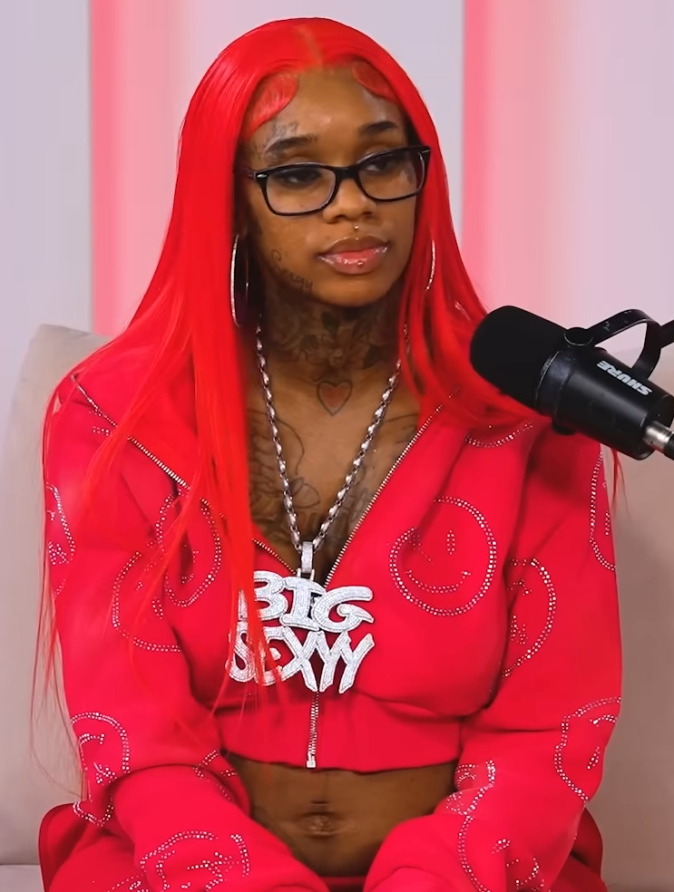 Sexyy Red sits at a podcast microphone in bright red hair and a matching red tracksuit, wearing black-framed glasses.