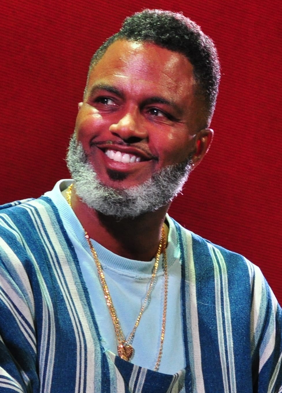 Ishmael Butler of Shabazz Palaces smiles while wearing gold chains and a striped cardigan over a light sweater.