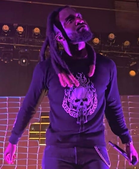 Shaboozey performs on stage under purple lighting, wearing a graphic sweatshirt with dreadlocks pulled back, holding a microphone.
