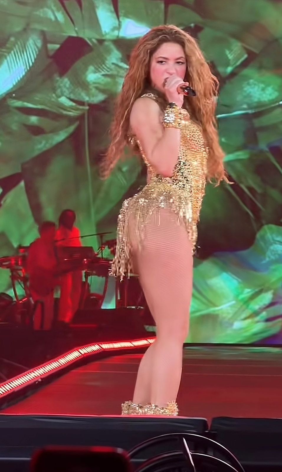 Shakira sings into a handheld microphone on a runway stage in a gold fringed bodysuit and gold boots, a green projection filling the screen behind her.