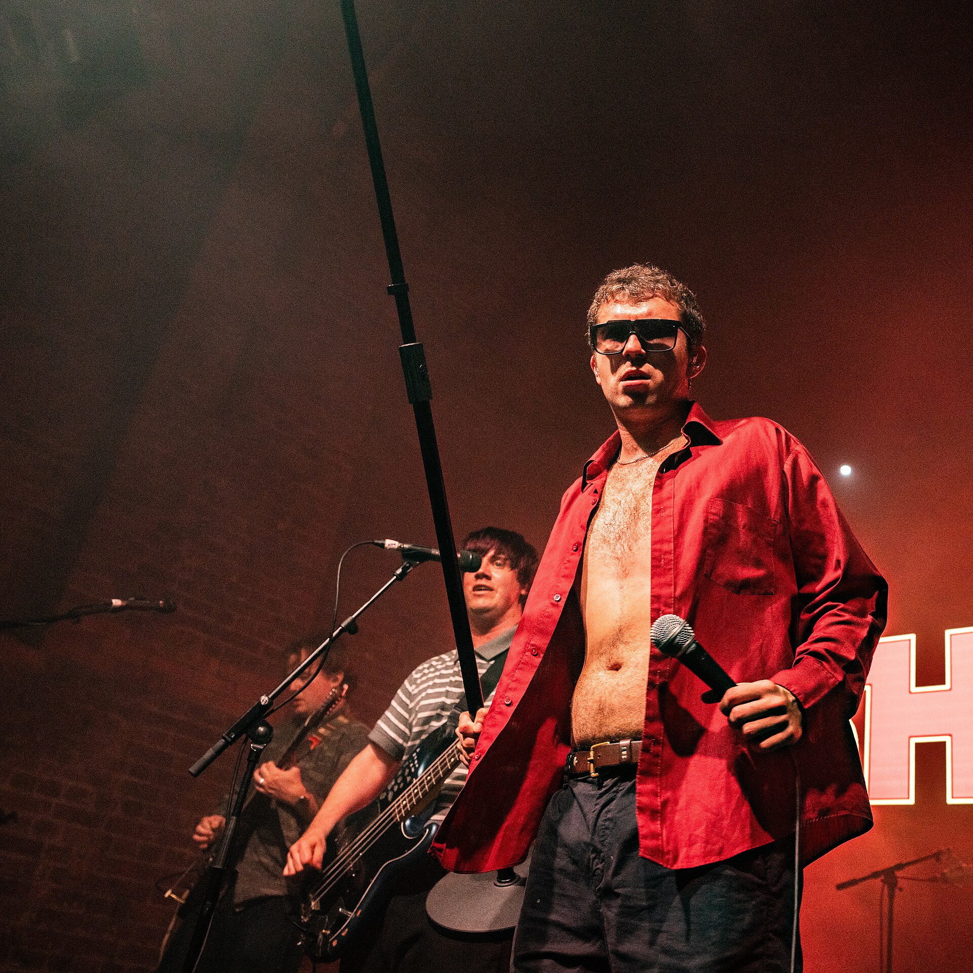 The singer of shame stands front of stage in dark sunglasses and an open red shirt, microphone at his side, the bassist and guitarist behind him and a lit band sign to the right.