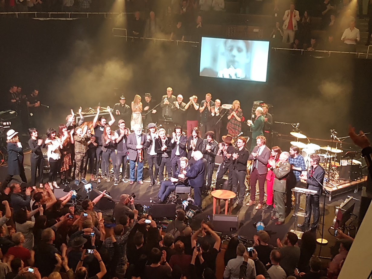 Seen from a balcony, a concert hall stage crowded with musicians applauds as a seated man is handed an award at the centre, a black and white portrait of him on the screen above.