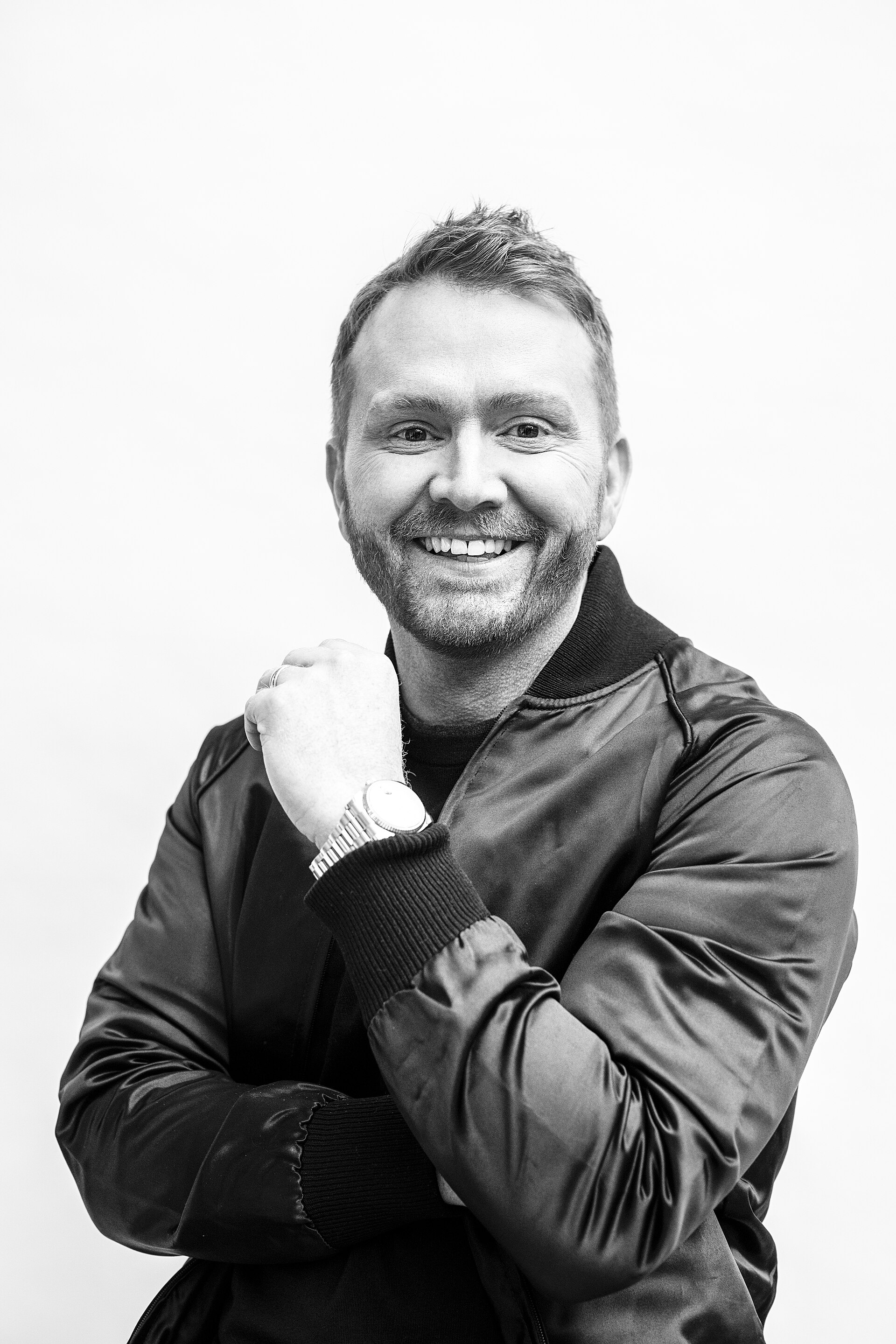 Black and white portrait of Shane McAnally smiling with his arms folded across his chest in a satin bomber jacket over a black sweater, against a plain pale background