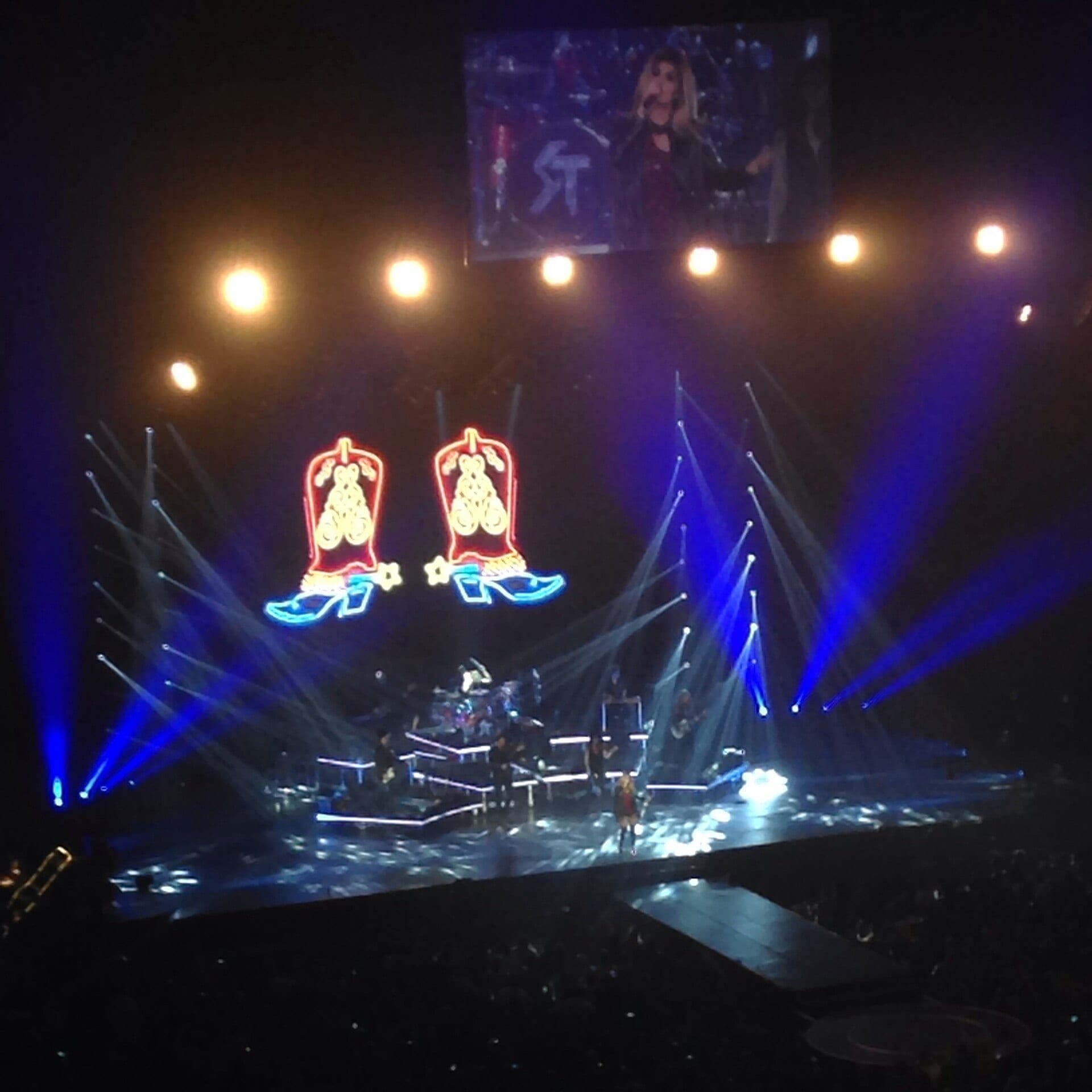 Shania Twain performs on an arena stage lit in blue beneath neon cowboy boot graphics, her face shown on the video screen above.