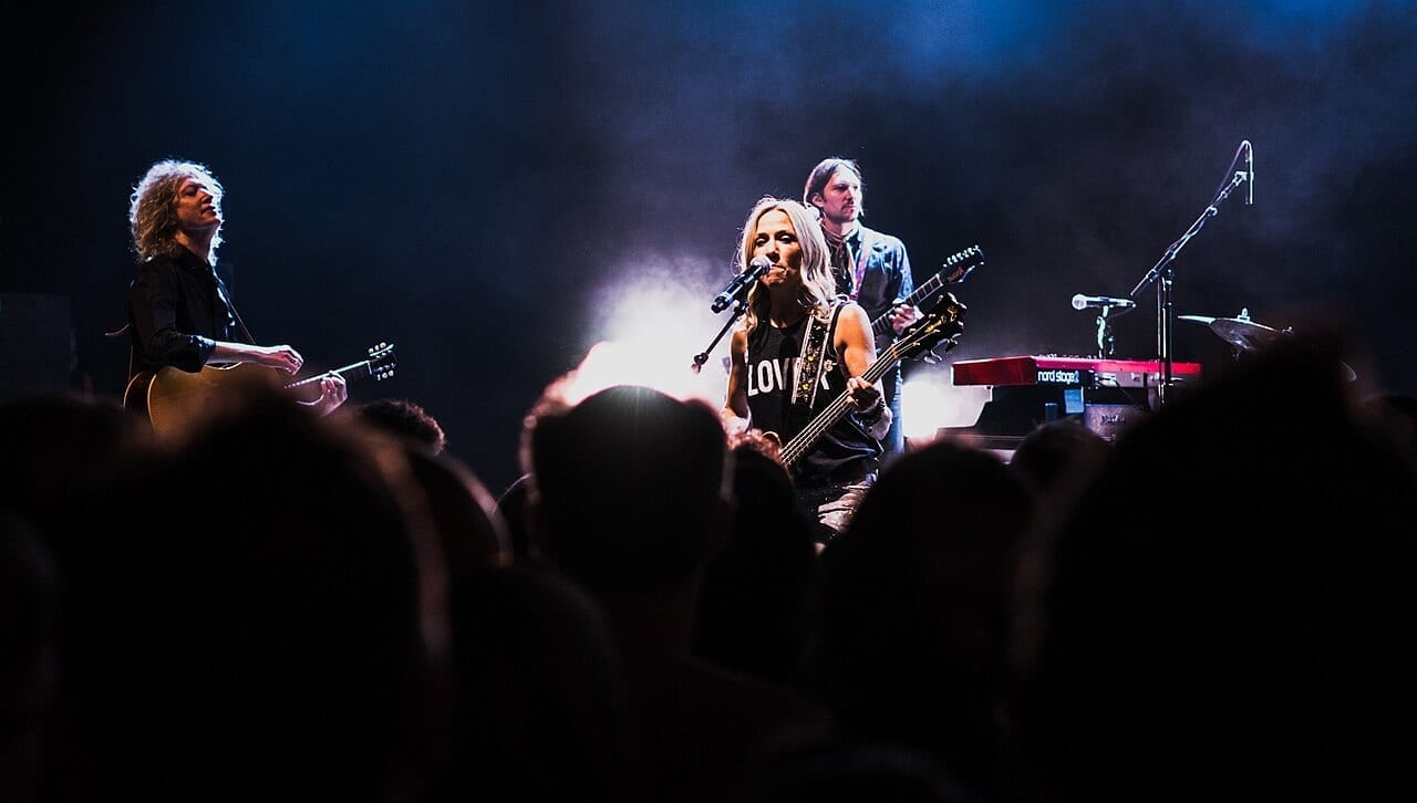 Sheryl Crow sings at a microphone while playing bass guitar, flanked by two band members on a smoky stage.