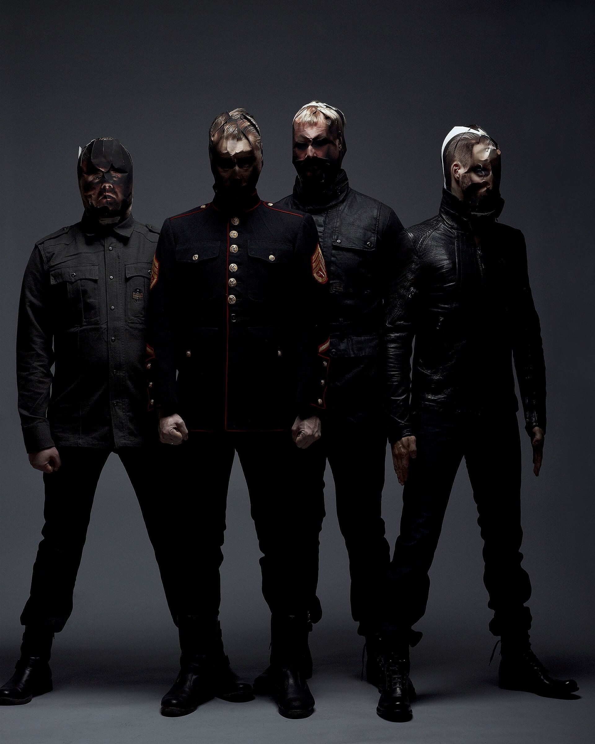 Studio portrait of the four masked members of Shining, dressed in black military-style jackets, standing in a row against a dark gray background.