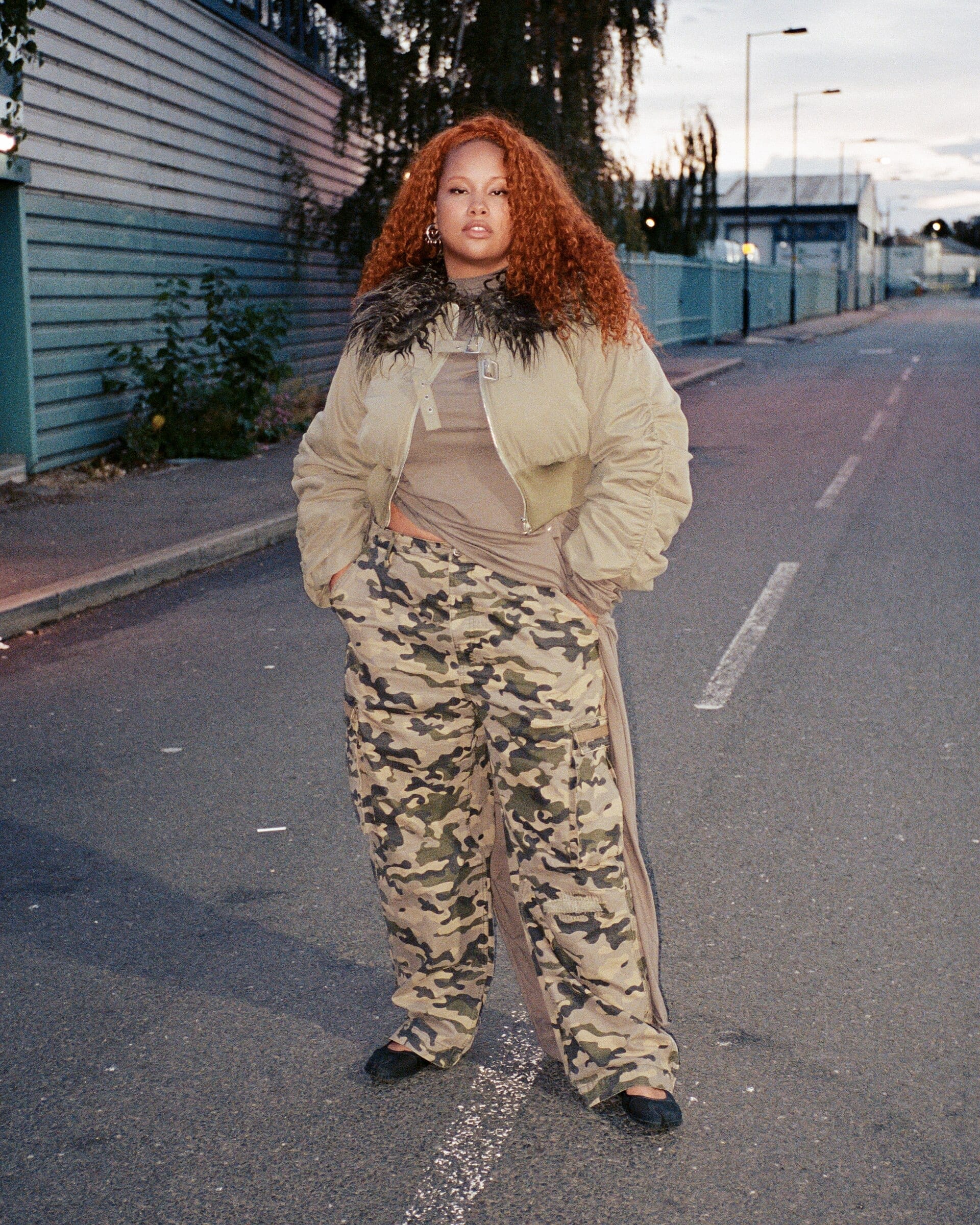 Shygirl stands in the middle of an empty road at dusk in a cream padded jacket with a black fur collar and camouflage cargo trousers, long ginger curls past her shoulders.