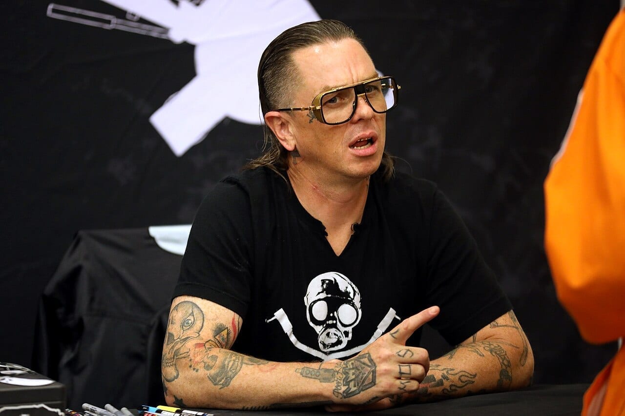 Sid Wilson of Slipknot, unmasked, sits at a signing table wearing tinted glasses and a black T-shirt printed with a gas-mask skull design, talking and gesturing with tattooed arms resting on the table.