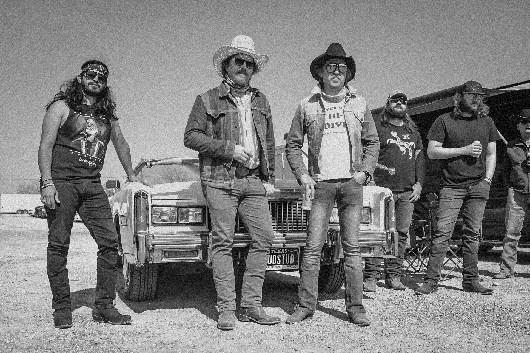 Five men in denim, cowboy hats and boots stand around the front of an old Cadillac on gravel, photographed in black and white