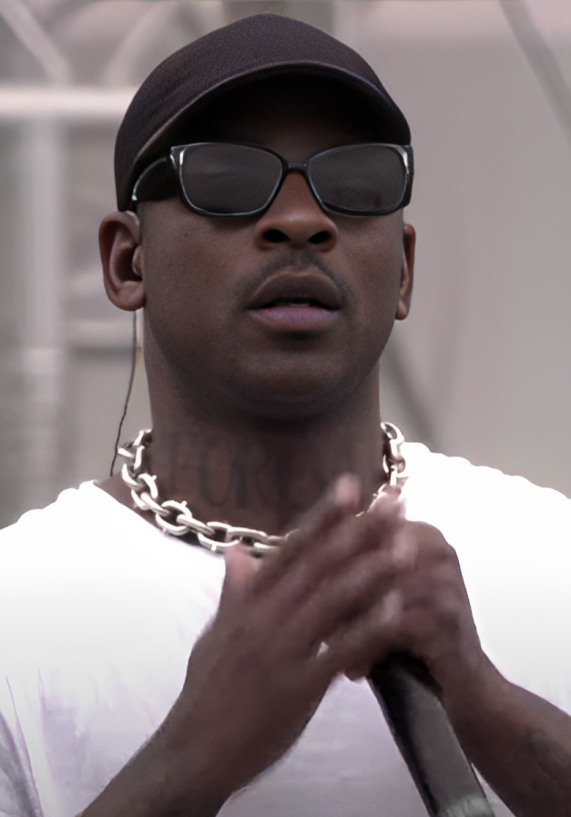 Skepta stands at a microphone stand in a black cap and sunglasses, a white T-shirt and a heavy silver chain, both hands on the mic.
