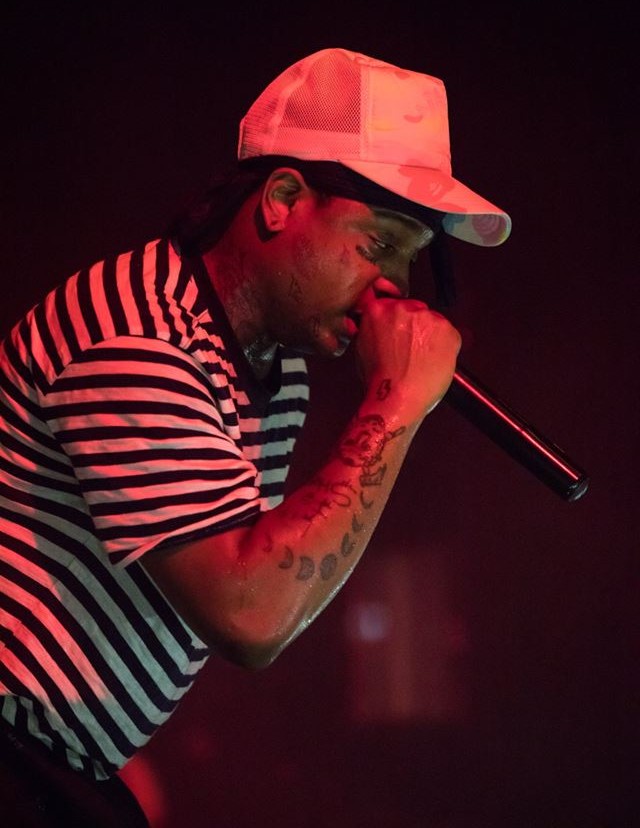 A rapper in a striped shirt and a mesh cap leans into a handheld microphone under red stage light, face and forearm tattoos visible