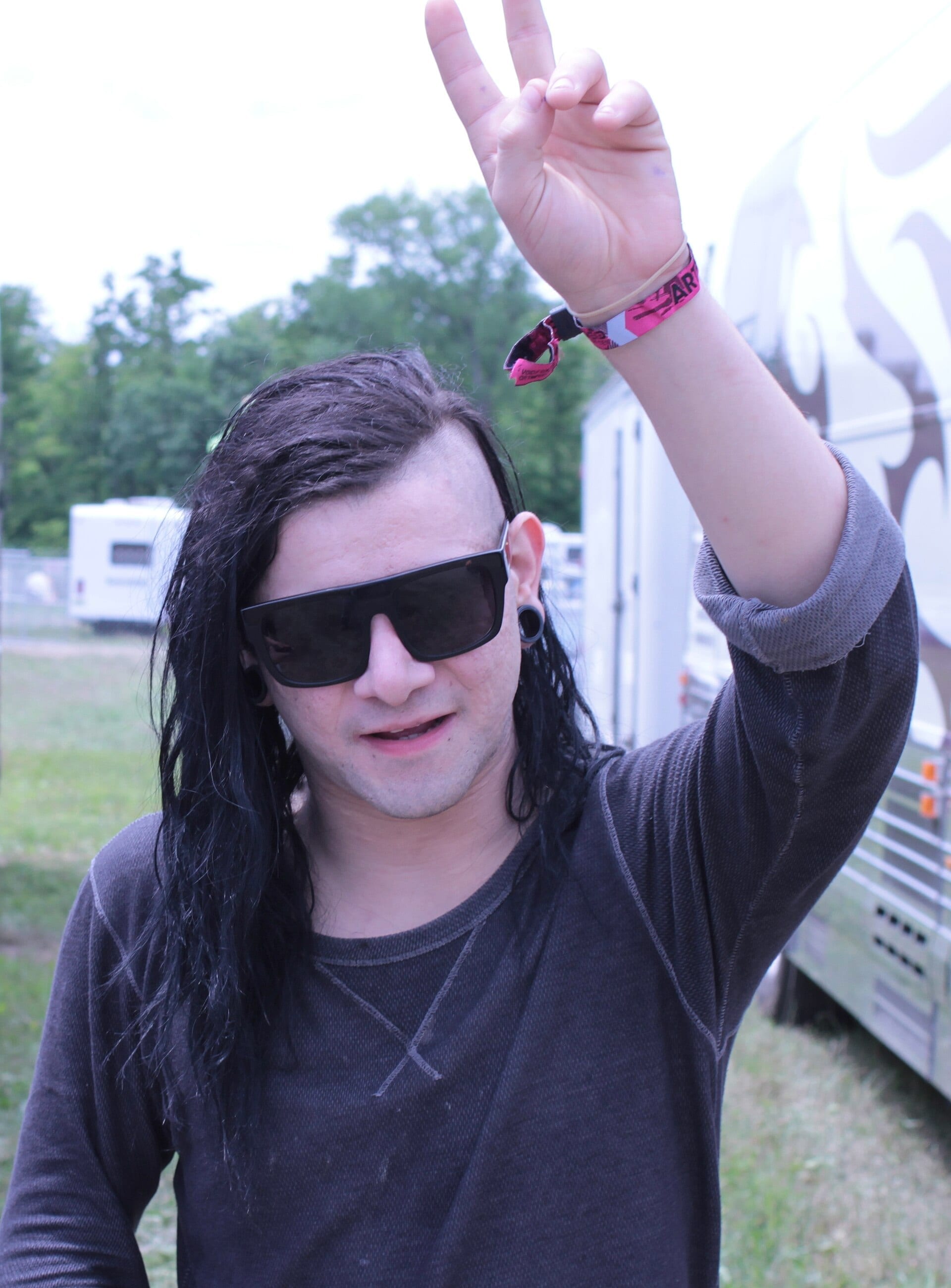 Skrillex stands outdoors near a tour bus, wearing dark sunglasses and a long-sleeve shirt, holding up a peace sign with his signature undercut hairstyle visible.