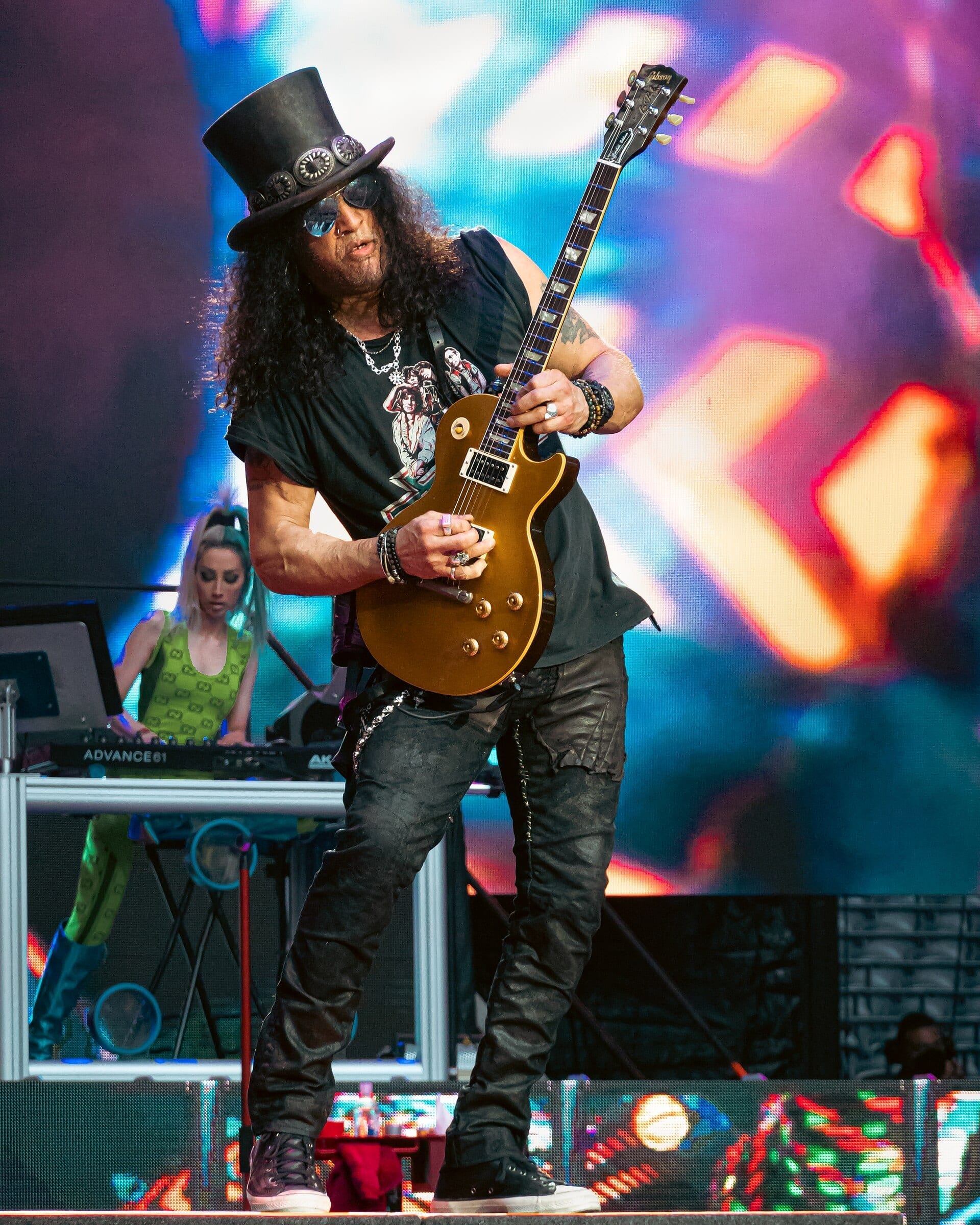 Slash plays a gold-top Les Paul guitar on stage, wearing his signature top hat and sunglasses with long curly hair.