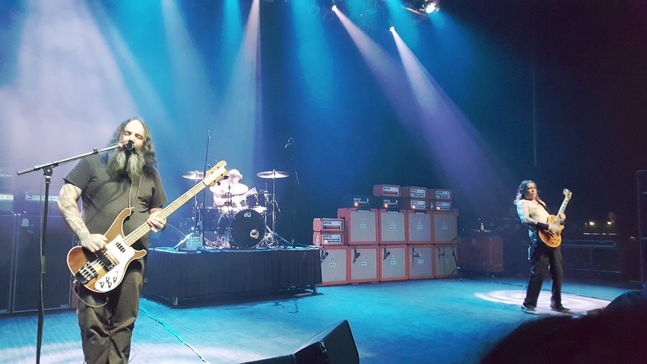 The band Sleep performing live under blue stage lighting: a bearded bassist singing into a microphone in the foreground, a drummer at a kit in the center background, and a guitarist playing at right, with stacked amplifier cabinets between them.