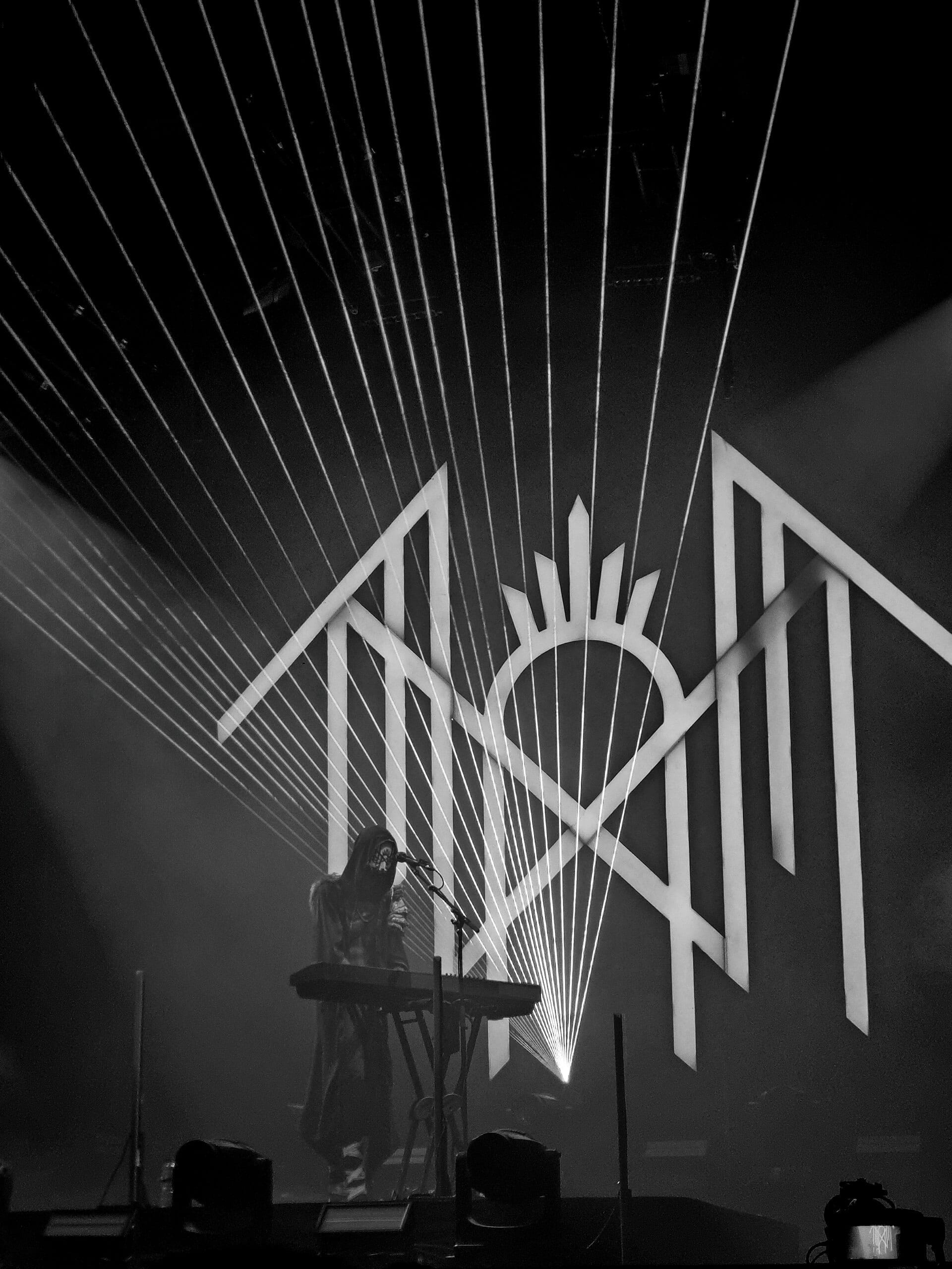A black-and-white arena shot of a masked, robed figure singing at a keyboard beneath Sleep Token's angular sigil, with laser lines fanning out across the ceiling.