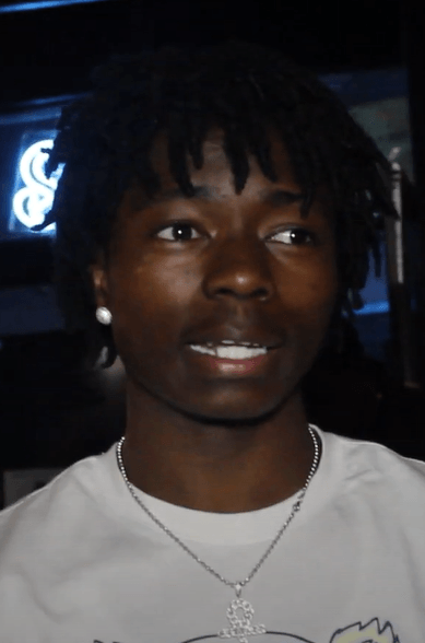 A close-up portrait of Sleepy Hallow, wearing a stud earring and a silver chain over a light gray t-shirt, with a neon sign glowing behind him.
