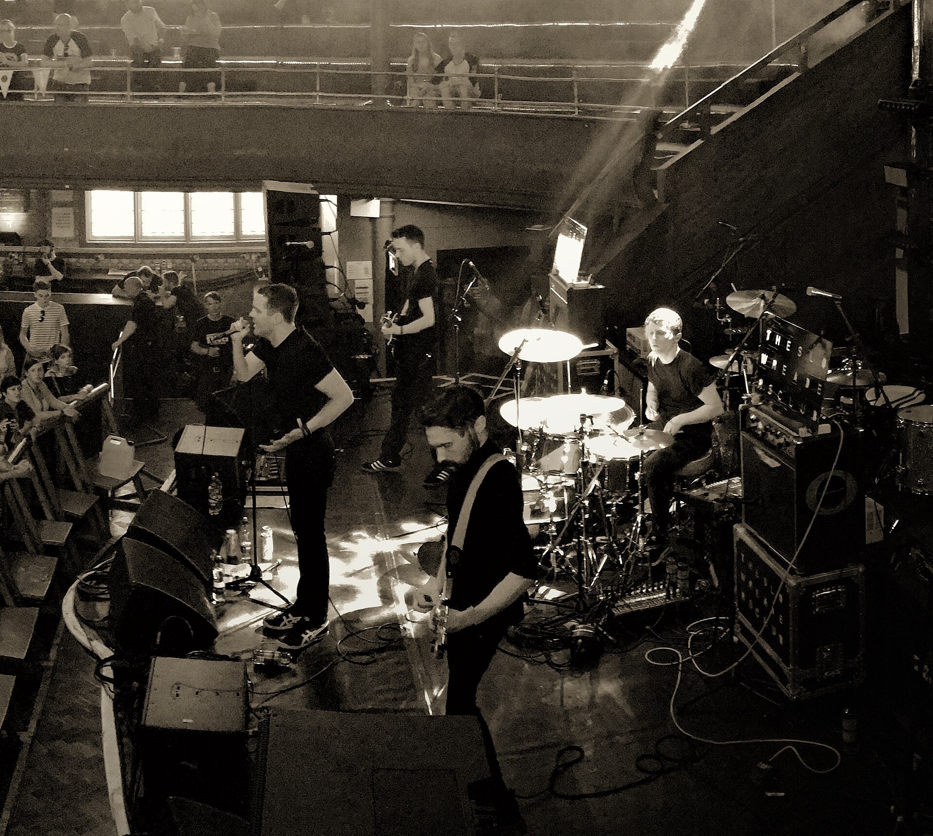 A four-piece band plays a low stage in a hall, shot from a balcony in warm sepia tones, with a small seated audience along one side.