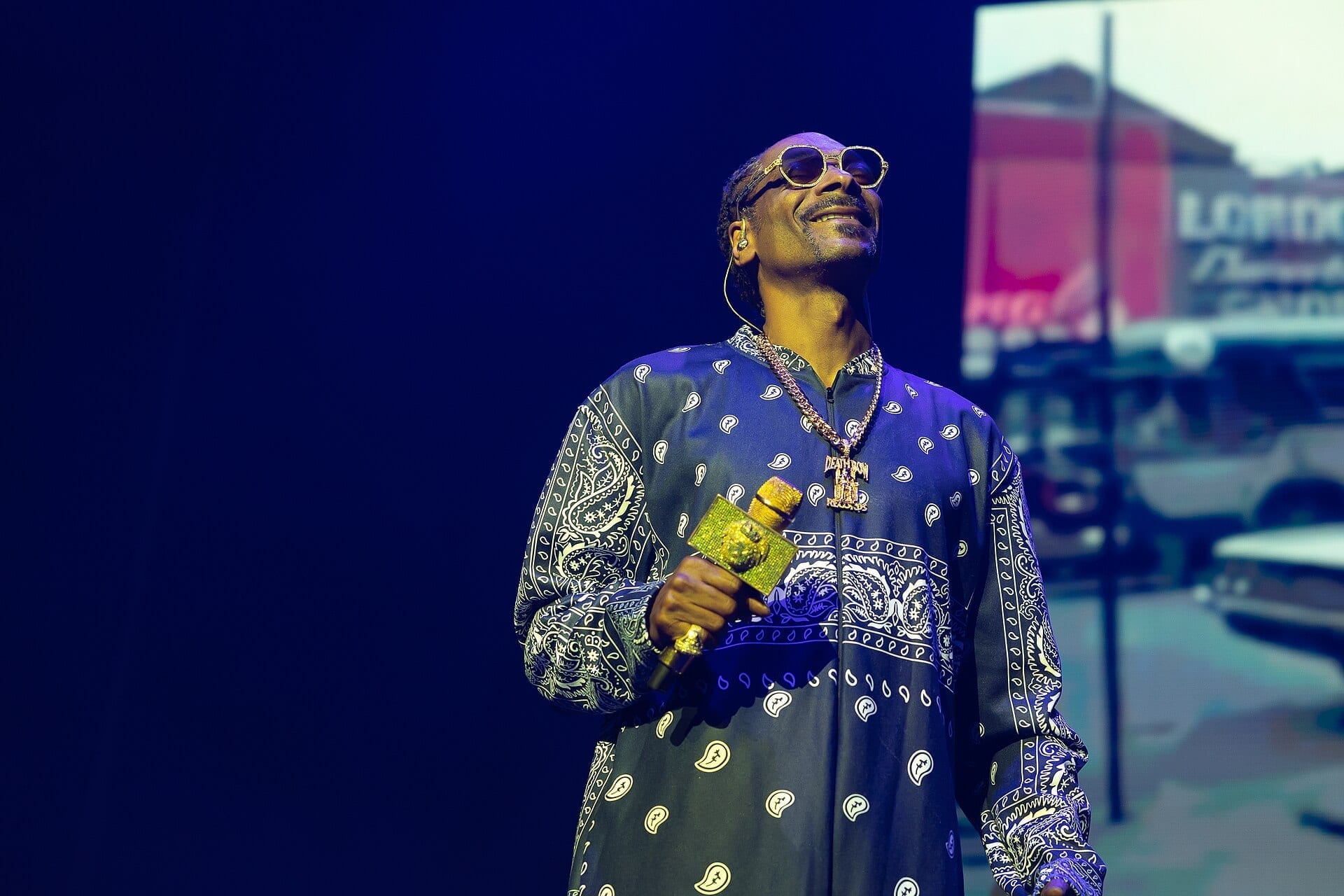 Snoop Dogg tips his head back and smiles on a dark stage, wearing gold-rimmed round sunglasses, a long bandana-print robe and a jewelled Death Row chain, holding a gem-covered microphone.