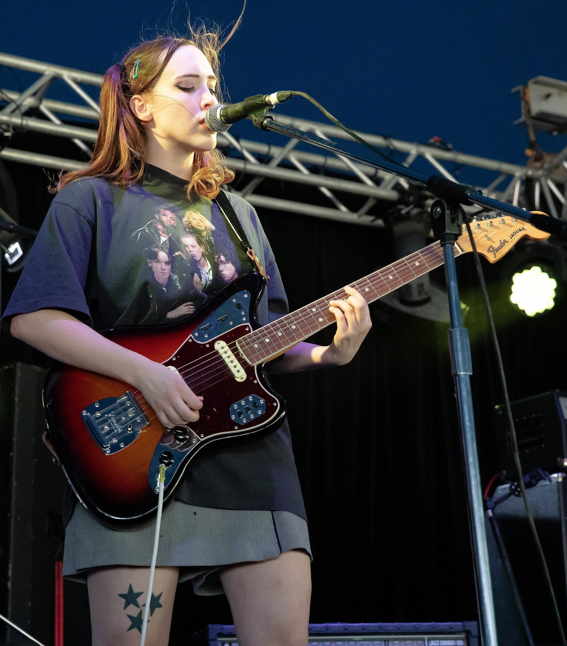 Sophie Allison of Soccer Mommy sings into a microphone while playing a sunburst Fender Jaguar guitar on an outdoor stage.