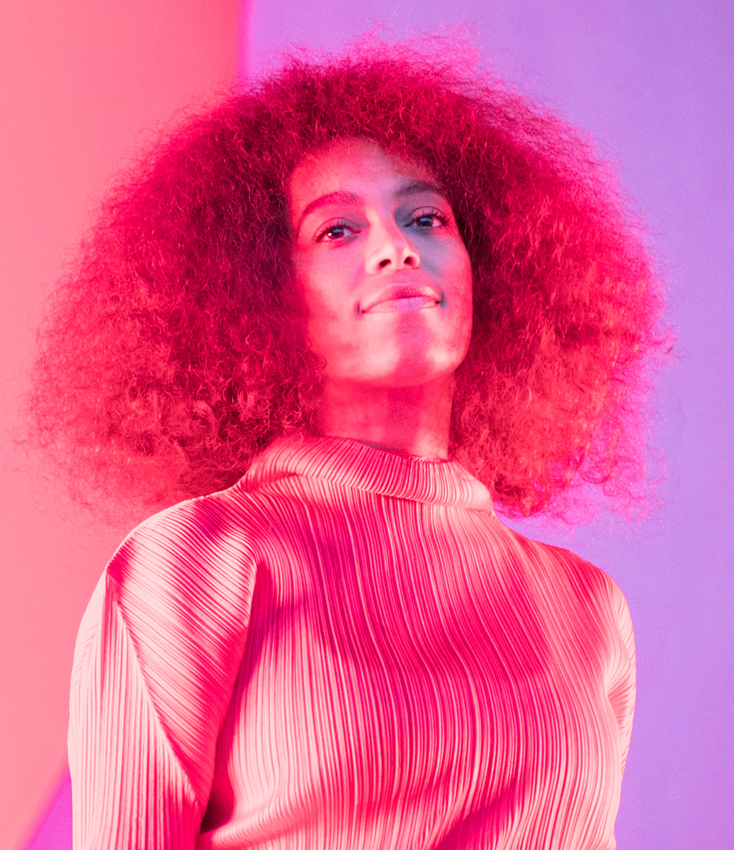 Solange on stage under pink and violet light, wearing a pleated high-necked top and looking slightly upward past the camera