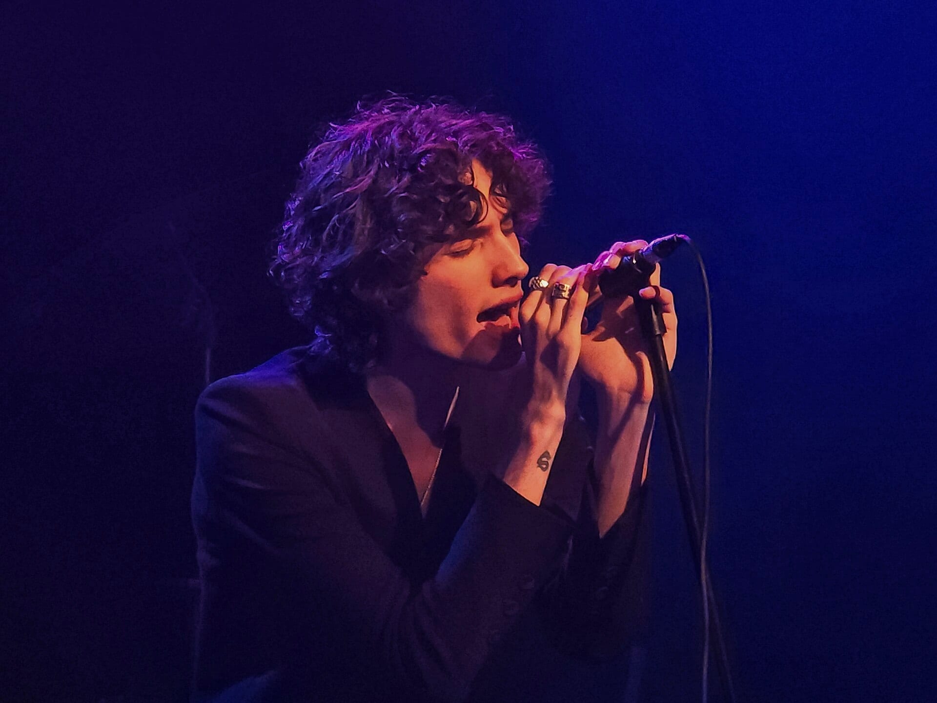 Sombr sings into a microphone on stage with his eyes closed, his curly hair lit purple and blue.