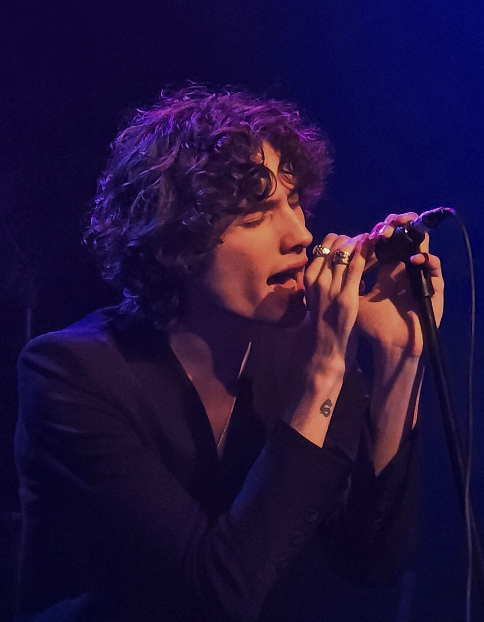 Sombr sings with his eyes closed into a mic held in both ringed hands, dark curly hair falling forward, lit purple against a blue backdrop.