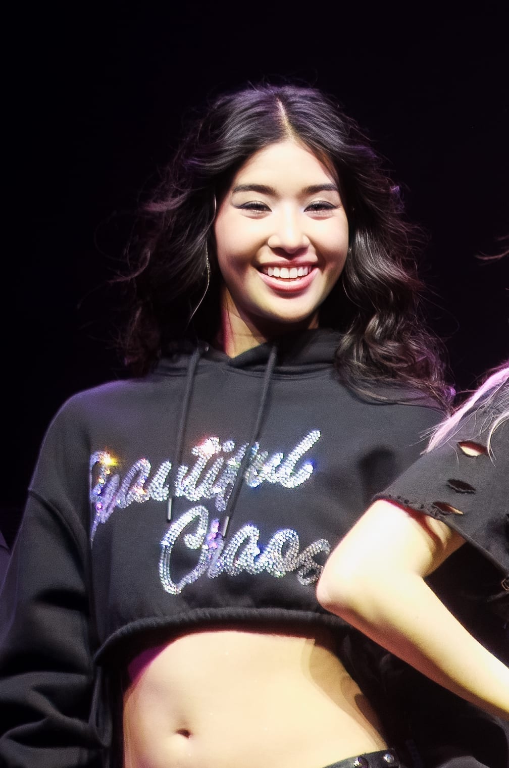 Sophia Laforteza smiles broadly on stage in a black cropped hoodie reading Beautiful Chaos, her dark hair falling loose.