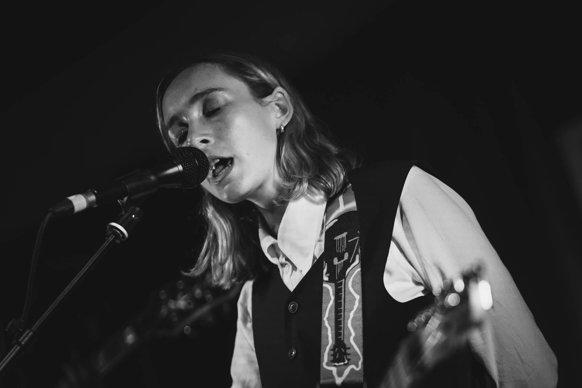 Black and white close-up of a singer with a guitar strap over one shoulder, eyes closed, singing into a microphone.