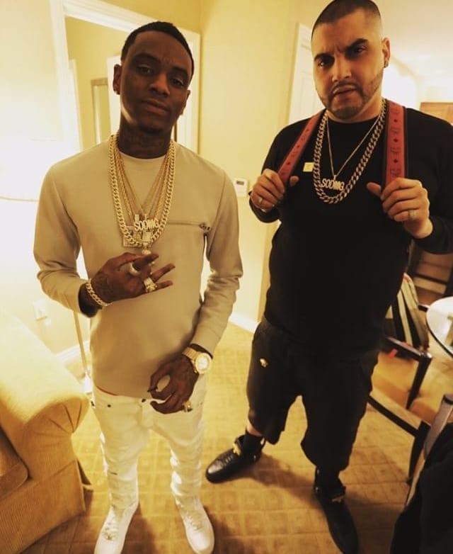 Soulja Boy poses indoors in layered gold chains and a cream sweater next to another man in a black shirt with a gold chain, both facing the camera.