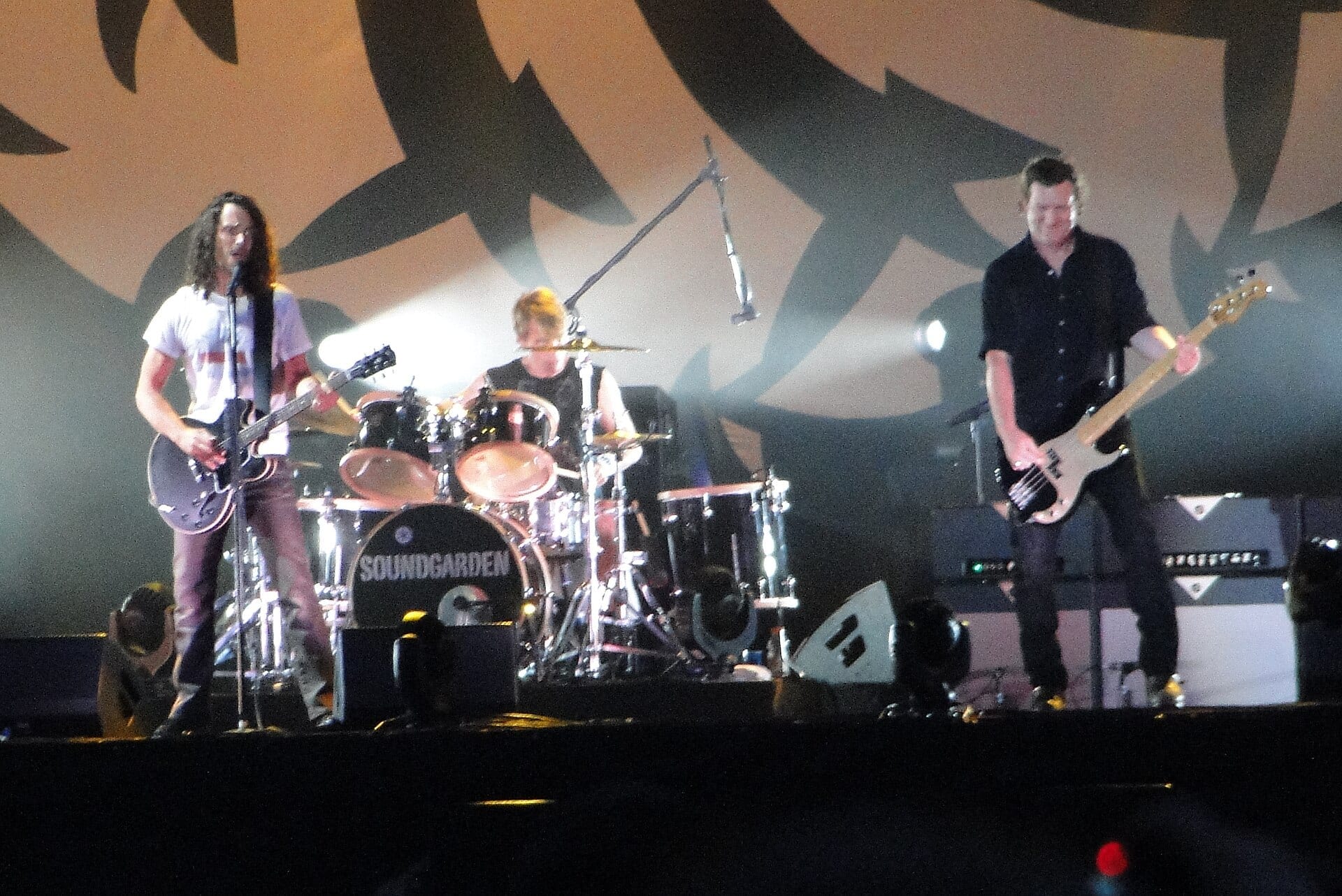 Three members of Soundgarden play a wide festival stage, a singer with a hollow-body guitar at left, a drummer behind a kit reading Soundgarden in the middle, and a bassist at right.