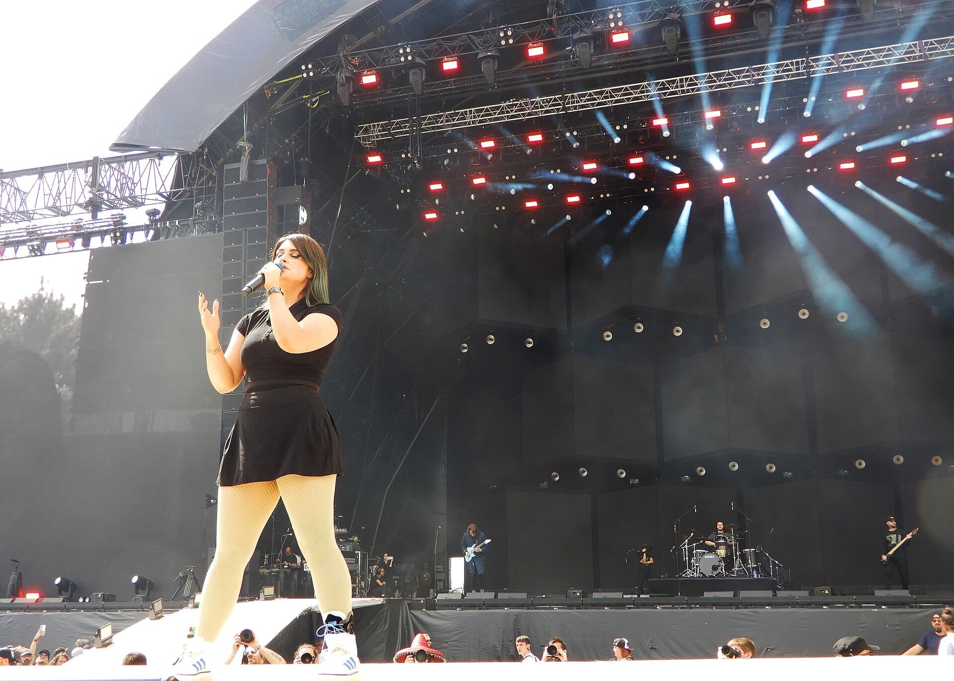 Courtney LaPlante sings into a handheld mic at the front of a daylit festival stage in a black top and skirt, green ends in her hair, her band spread out behind her.
