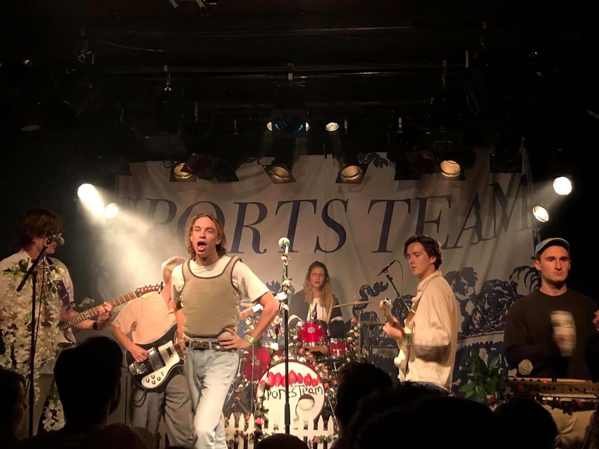Sports Team play a small club stage dressed with a white picket fence and flowers, the singer mid-shout in front of a banner bearing the band's name.