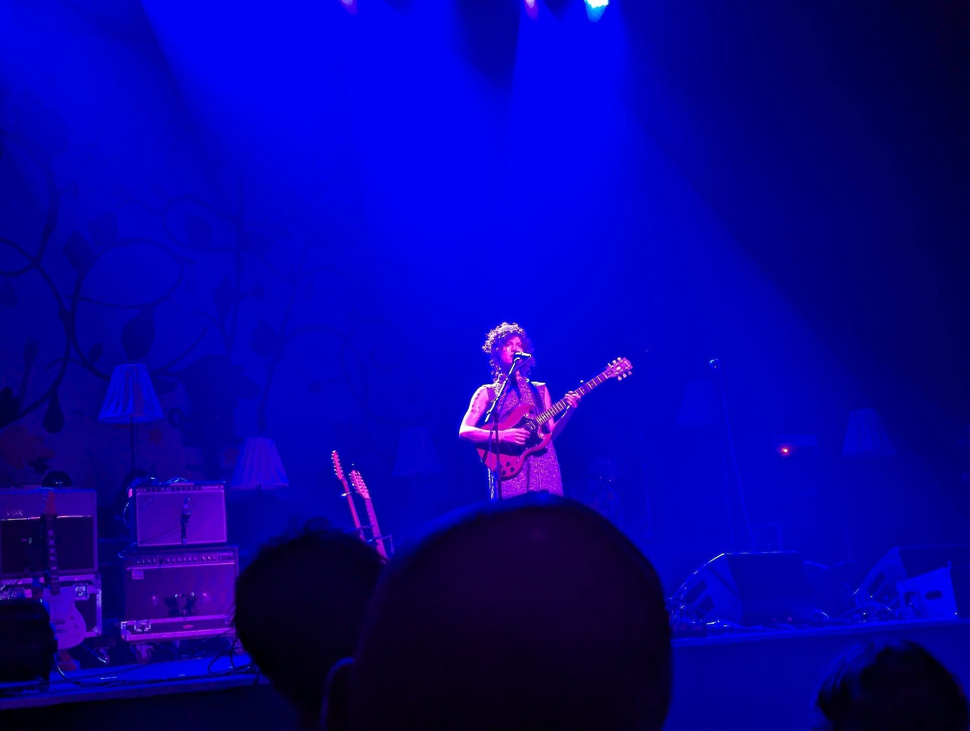 Squirrel Flower, with curly hair, sings into a microphone while playing an electric guitar, drenched in deep blue stage light.