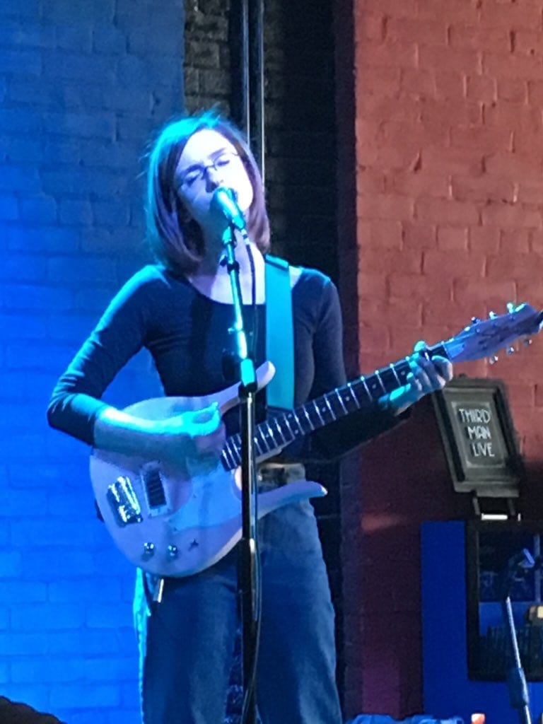 Stef Chura sings into a microphone with her eyes closed while playing an electric guitar with a teal strap, lit blue against a brick wall.