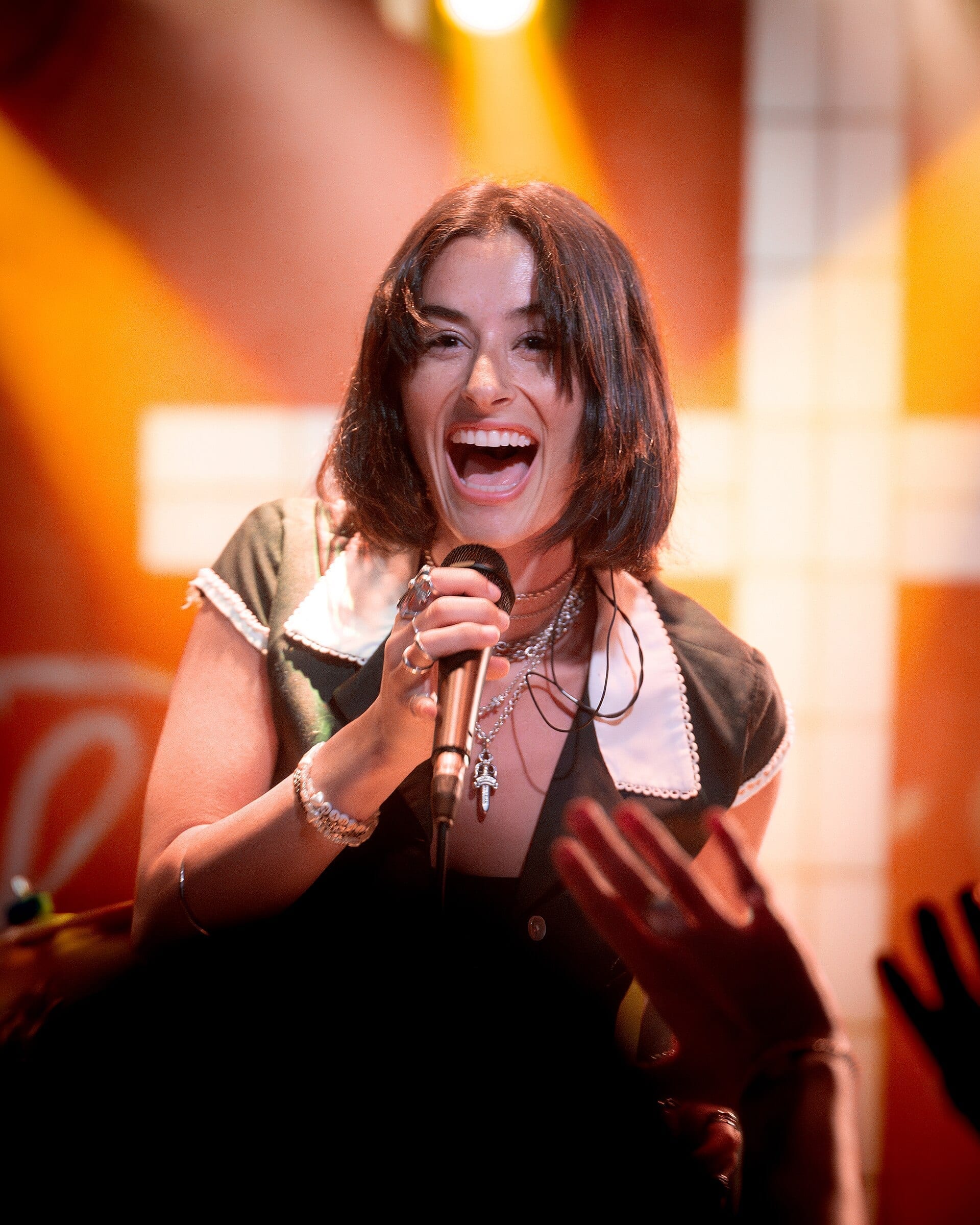 Stella Lefty singing into a microphone with an animated, joyful expression on stage, lit by warm orange stage lighting.