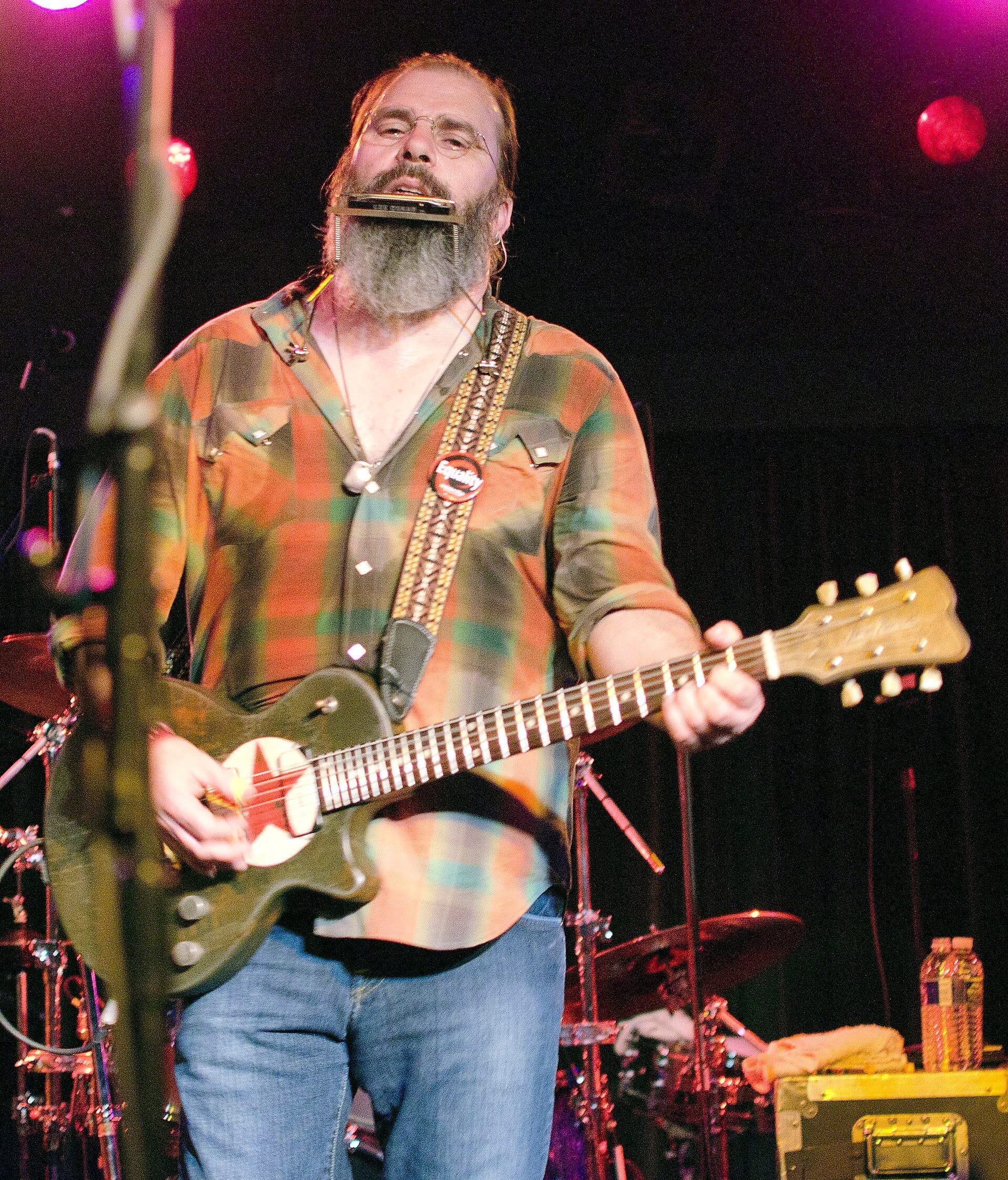 Steve Earle plays a harmonica mounted on a neck rack while strumming an olive-green electric guitar on stage, wearing a plaid shirt, an Equality button pin, and a full gray beard.