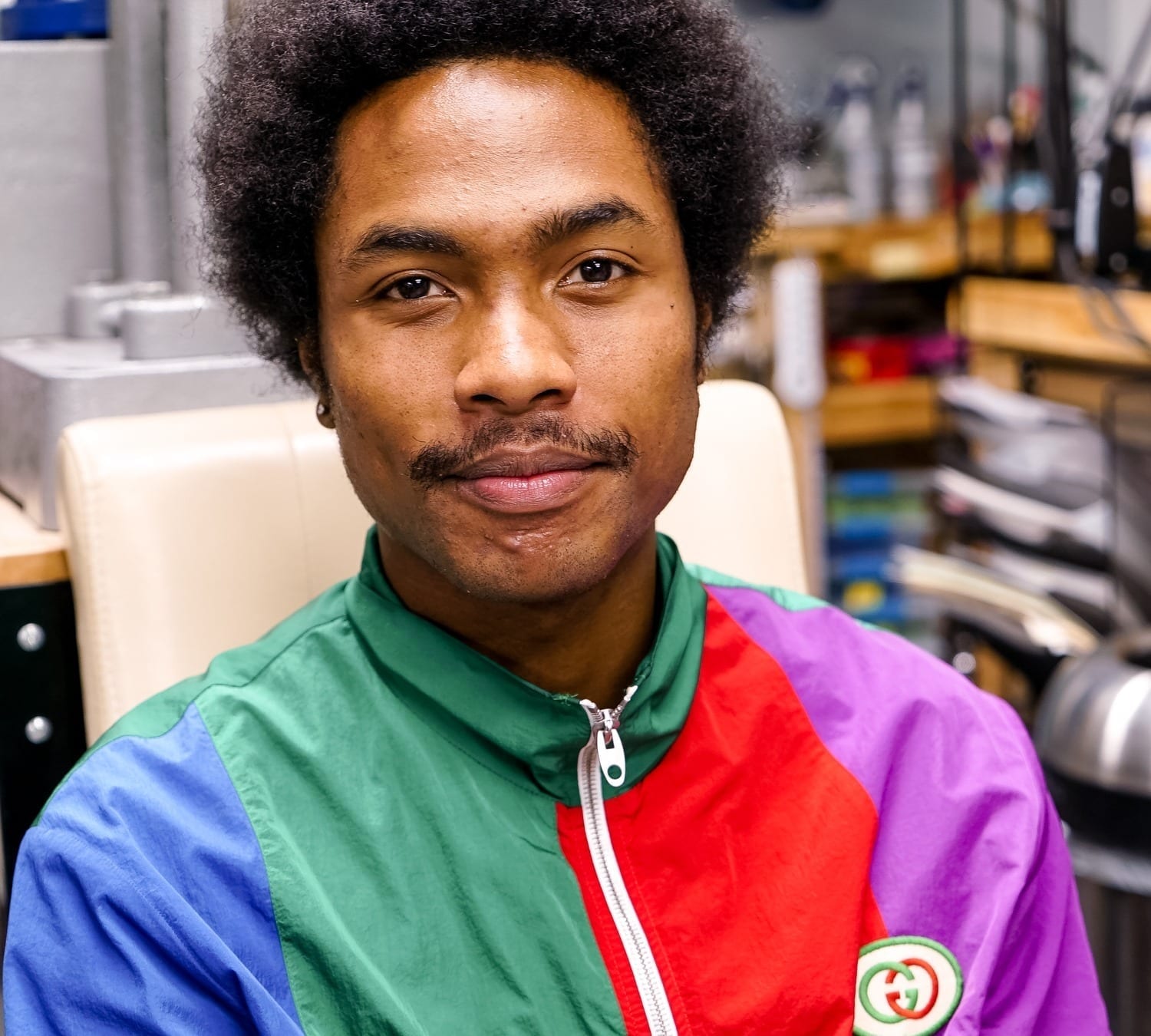 A close-up headshot of Steve Lacy, wearing a colorblocked windbreaker jacket, with a mustache and afro.