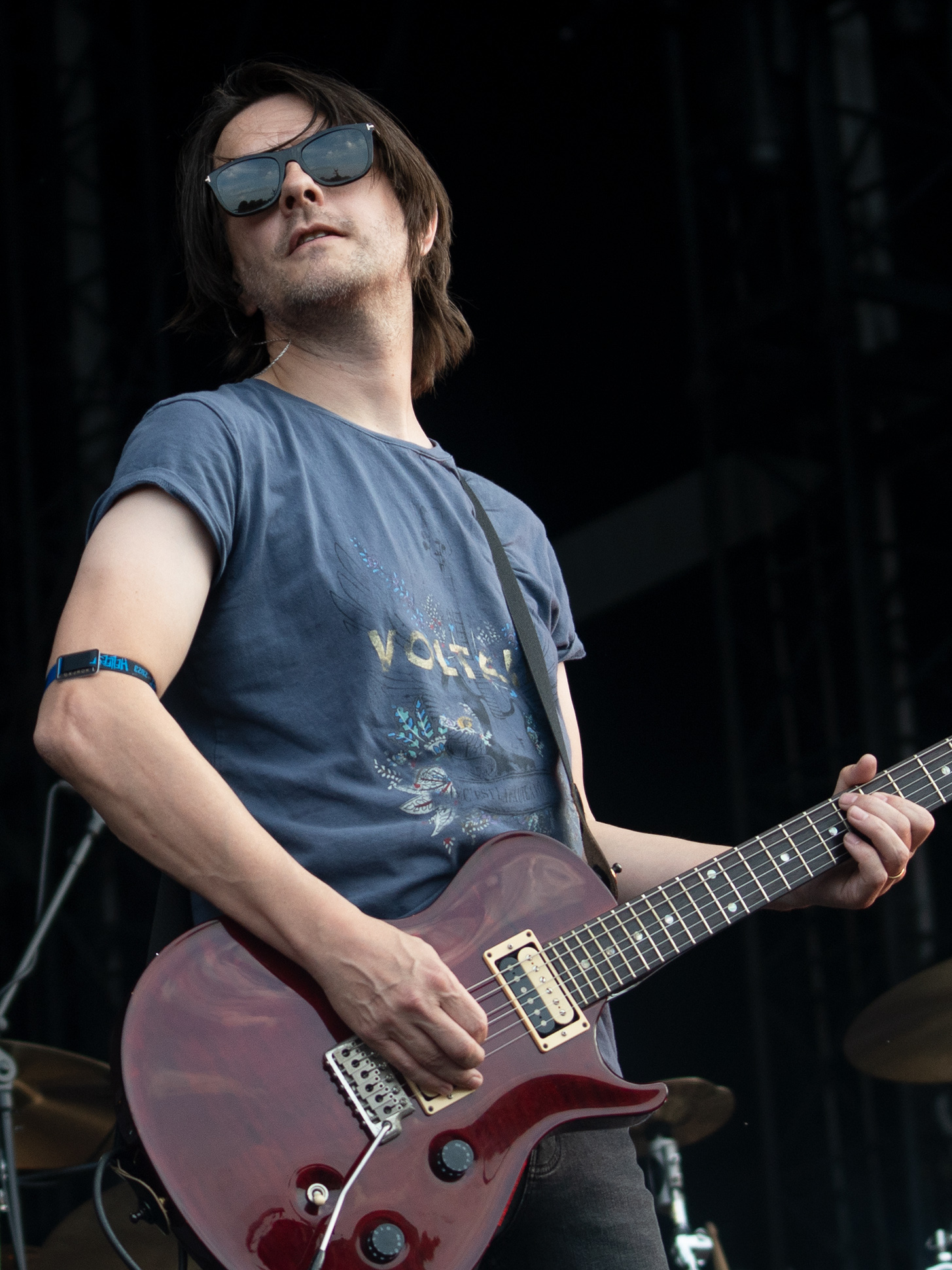 Steven Wilson playing a deep red electric guitar on an outdoor stage in sunglasses and a faded blue t-shirt, shoulder-length hair pushed back