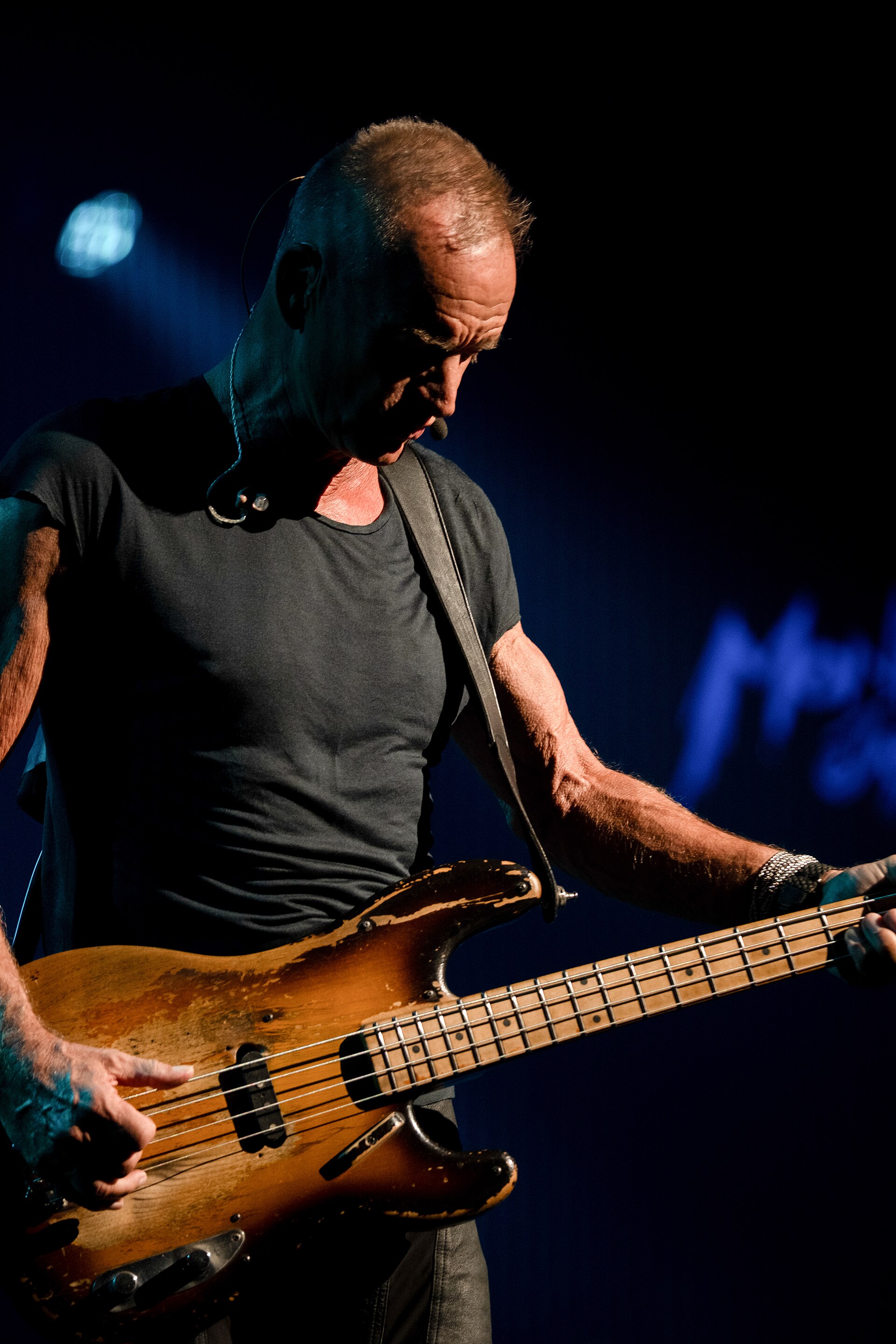 Sting looks down at the neck of a heavily worn sunburst bass guitar, in a grey T-shirt with a headset microphone.