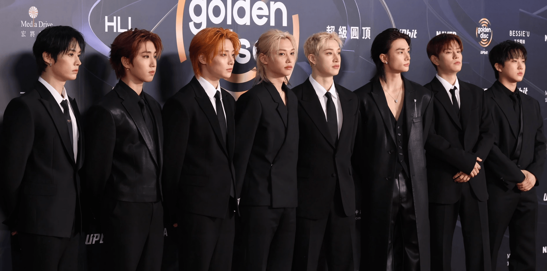 The eight members of Stray Kids stand in a line in black suits and ties on the Golden Disc Awards red carpet.