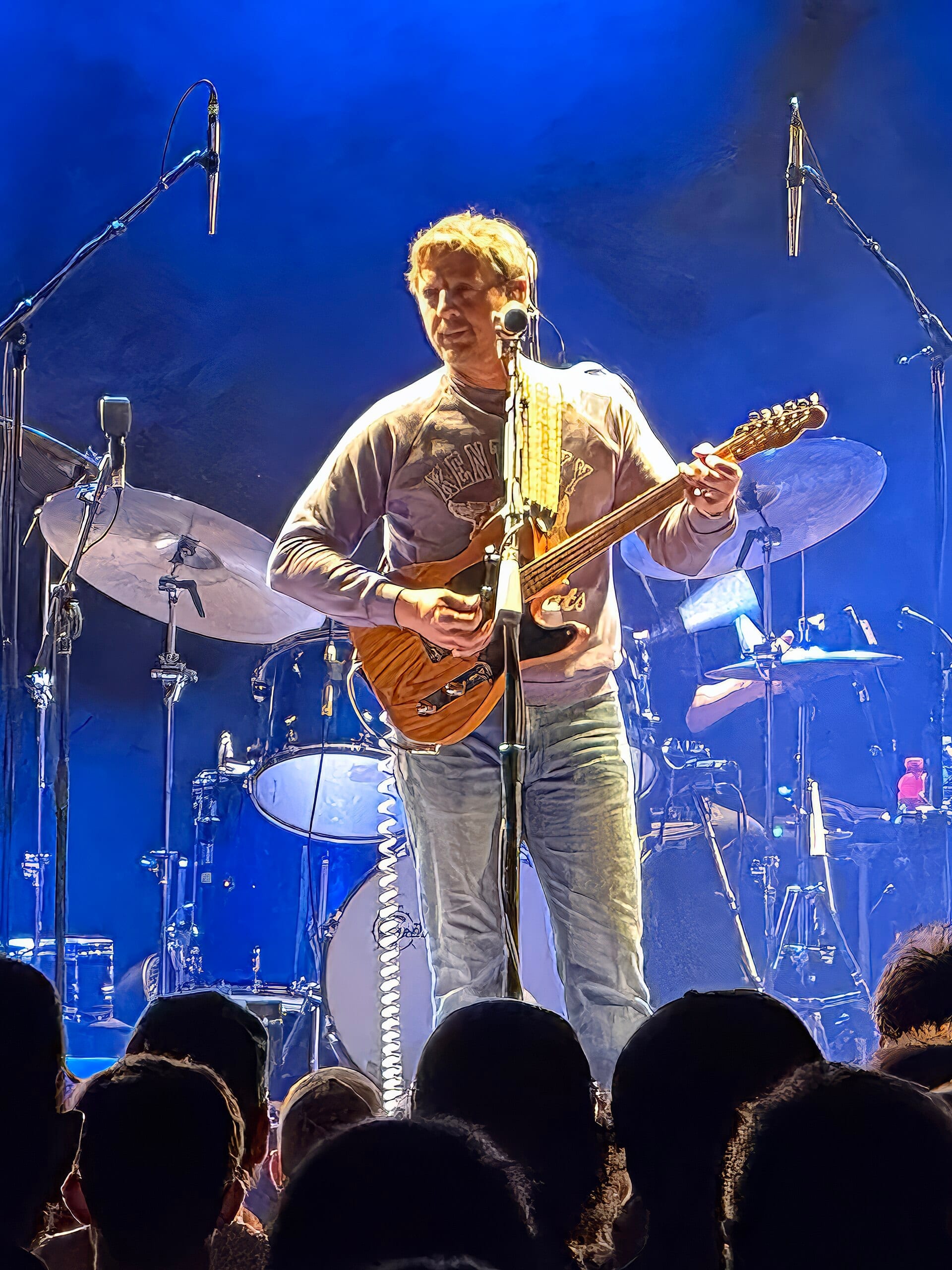 Sturgill Simpson stands at a microphone in a Kentucky sweatshirt and jeans, playing electric guitar in front of a blue-lit drum kit, heads of the front row in silhouette below.